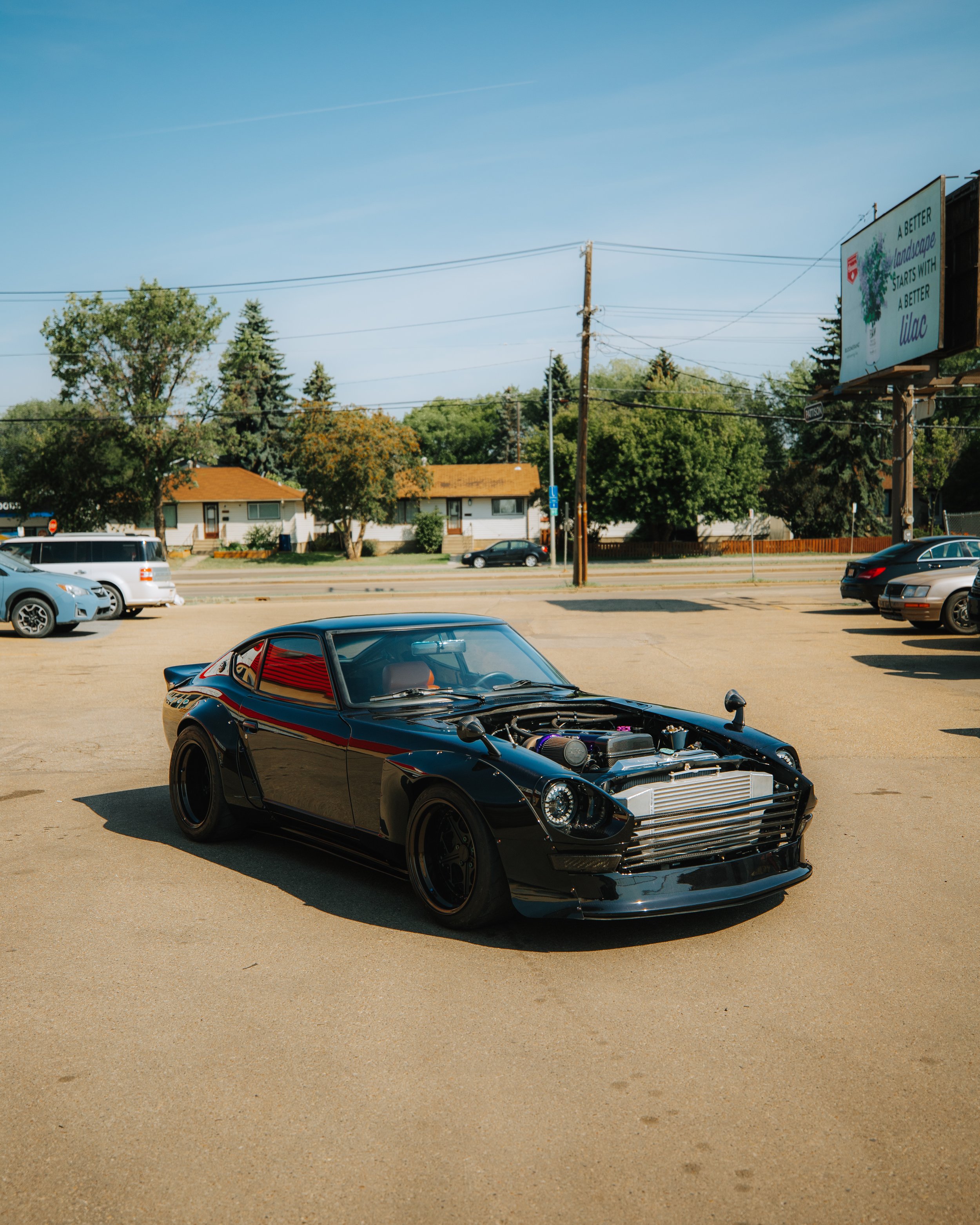 Black sports car with a modified front grille and exposed engine in a parking lot with other vehicles, houses, trees, and billboards in the background.