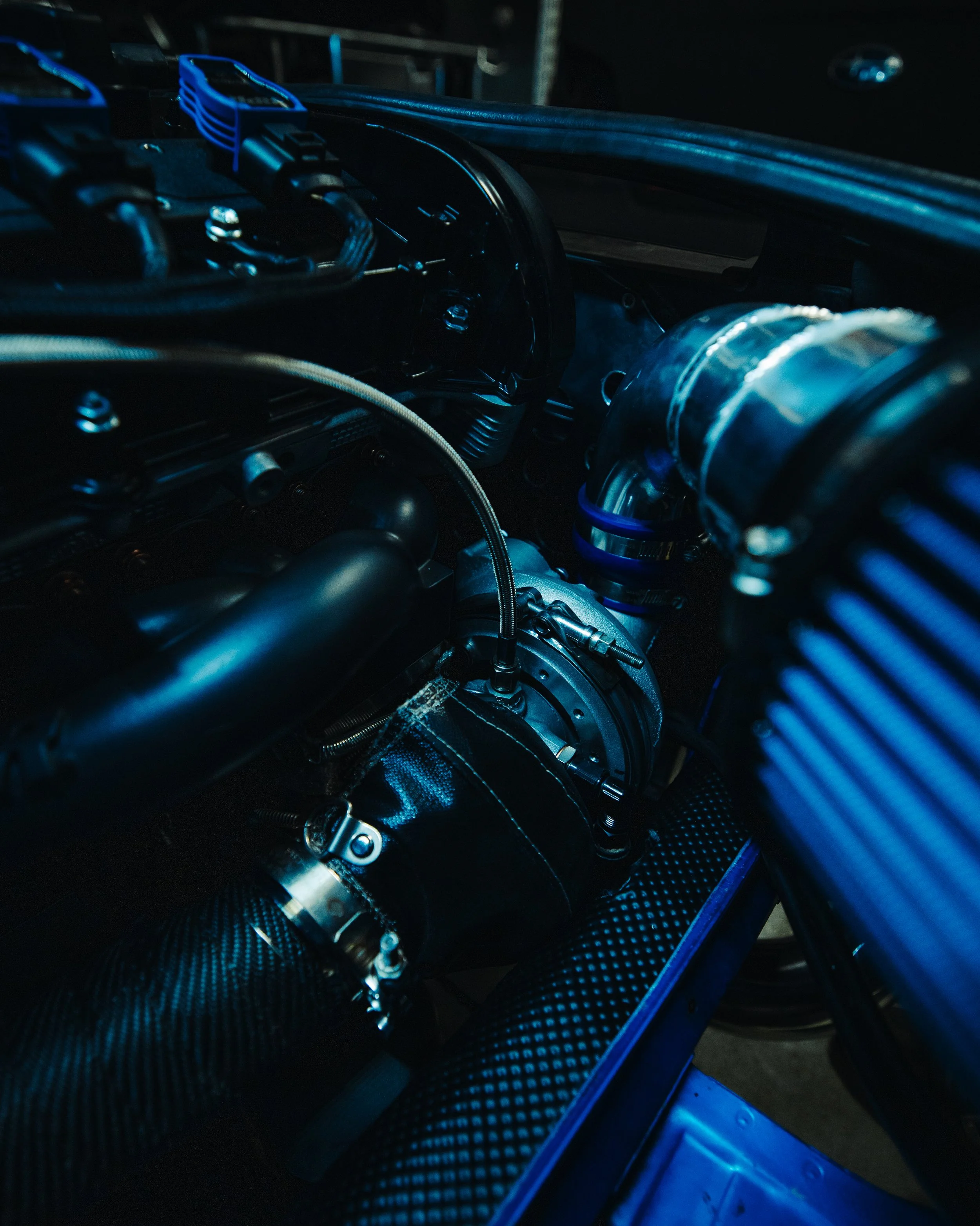Close-up of a high-performance car engine with various metal and plastic components, hoses, and connectors, illuminated with blue lighting.