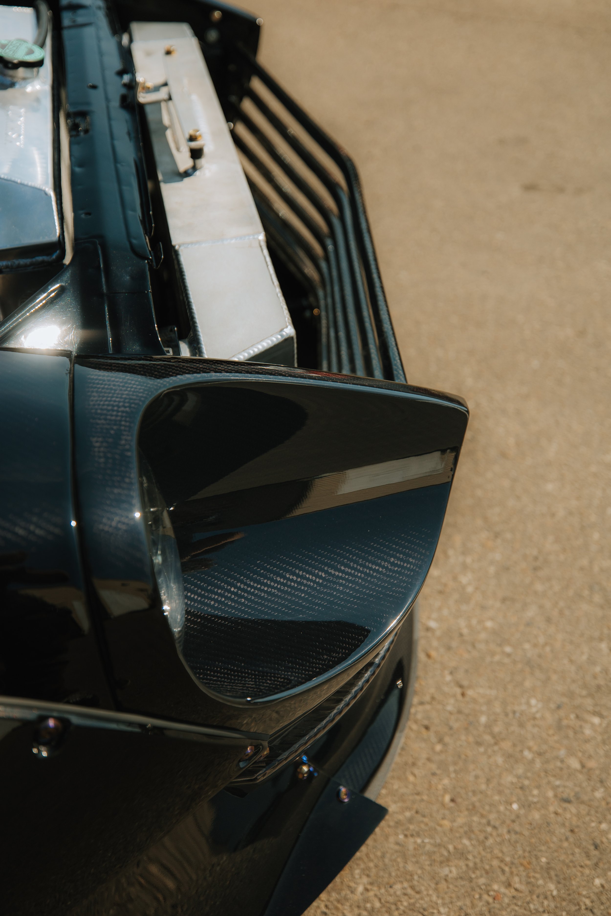 Close-up of the front right corner of a vintage black car showing the headlight, part of the grill, and the fender with a visible carbon fiber pattern.