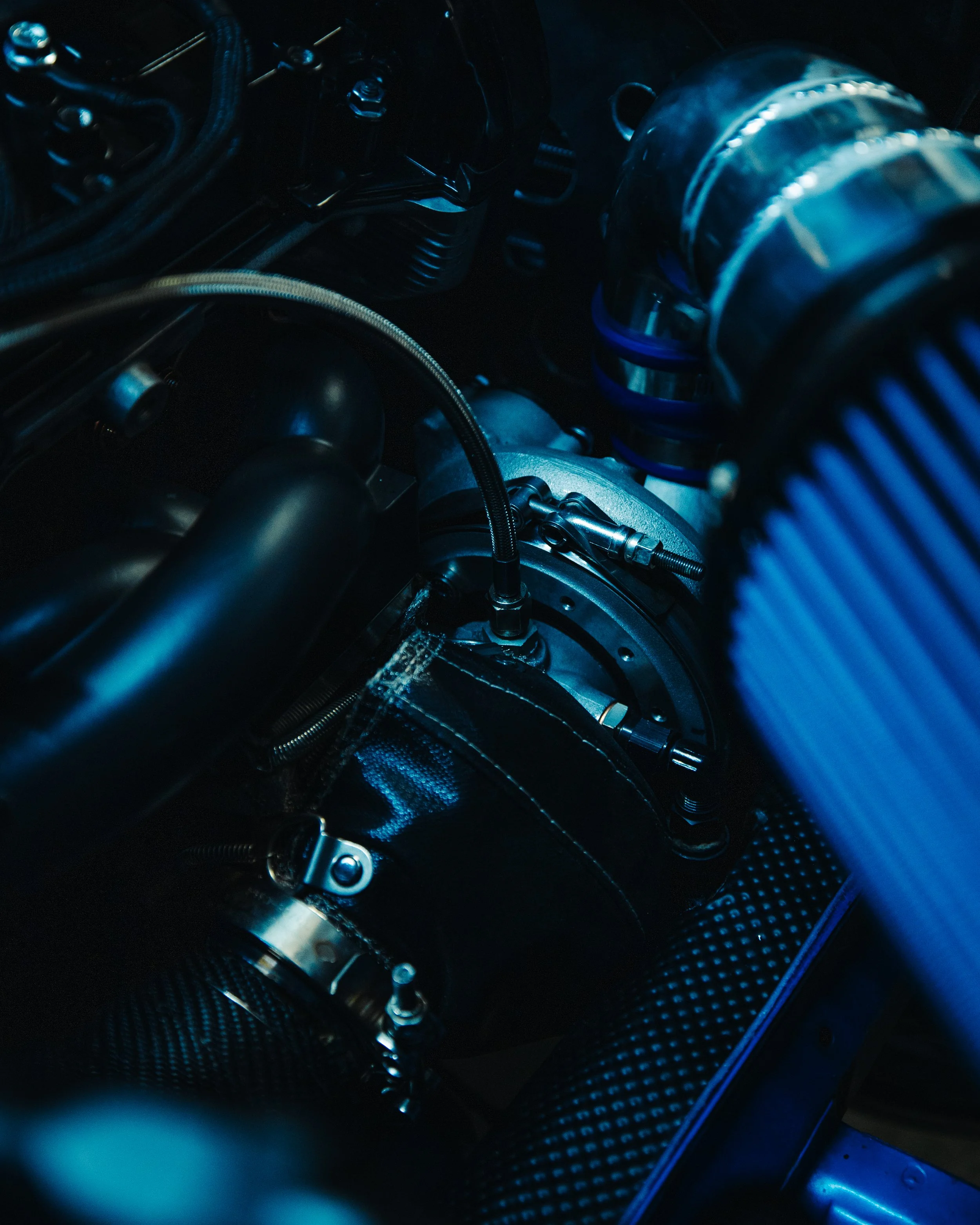 Close-up of a high-performance car engine with visible hoses, metal components, and a coil spring, illuminated with blue lighting.