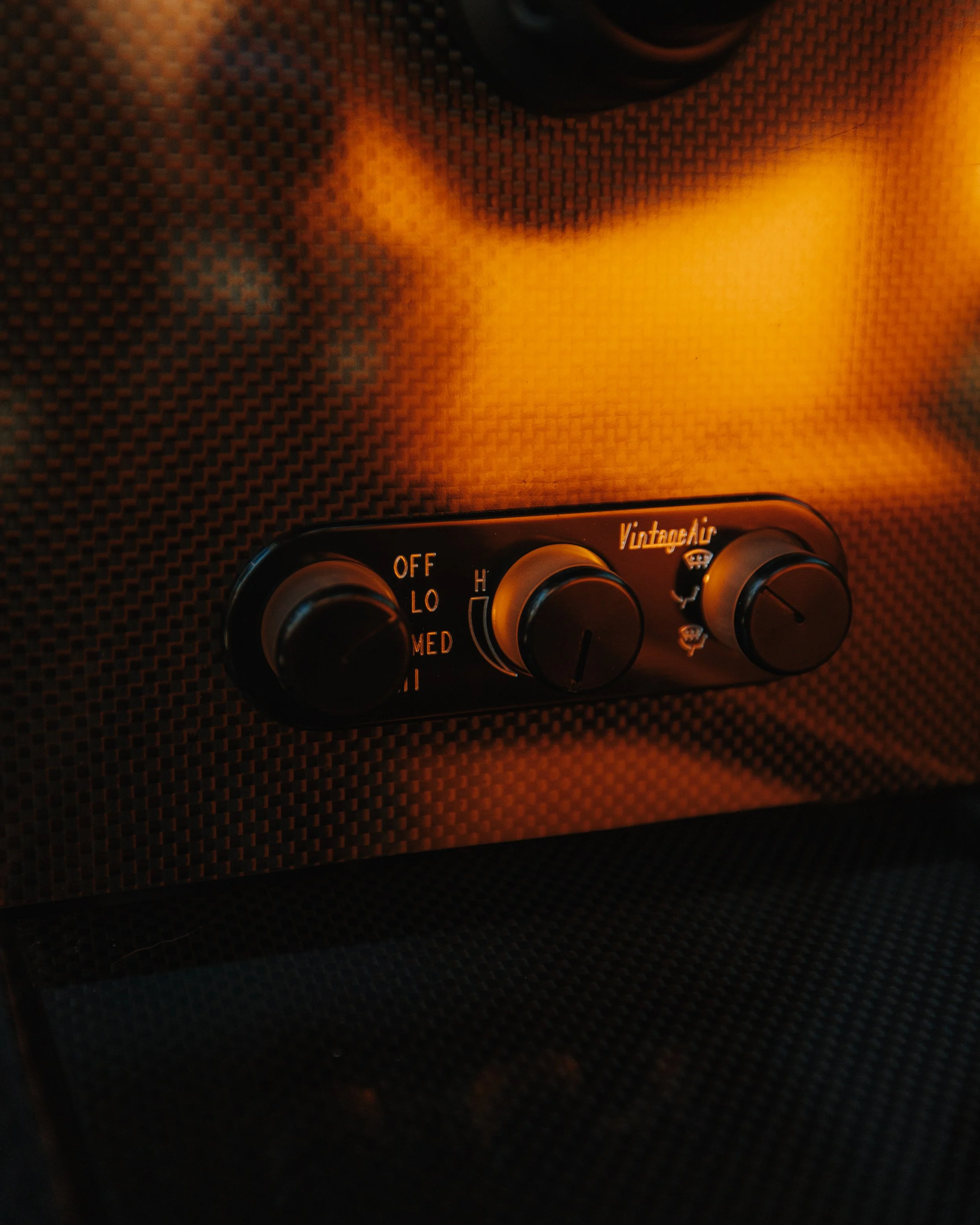 Close-up of a vintage car heater control panel with three black knobs, set on a textured black panel with orange lighting.