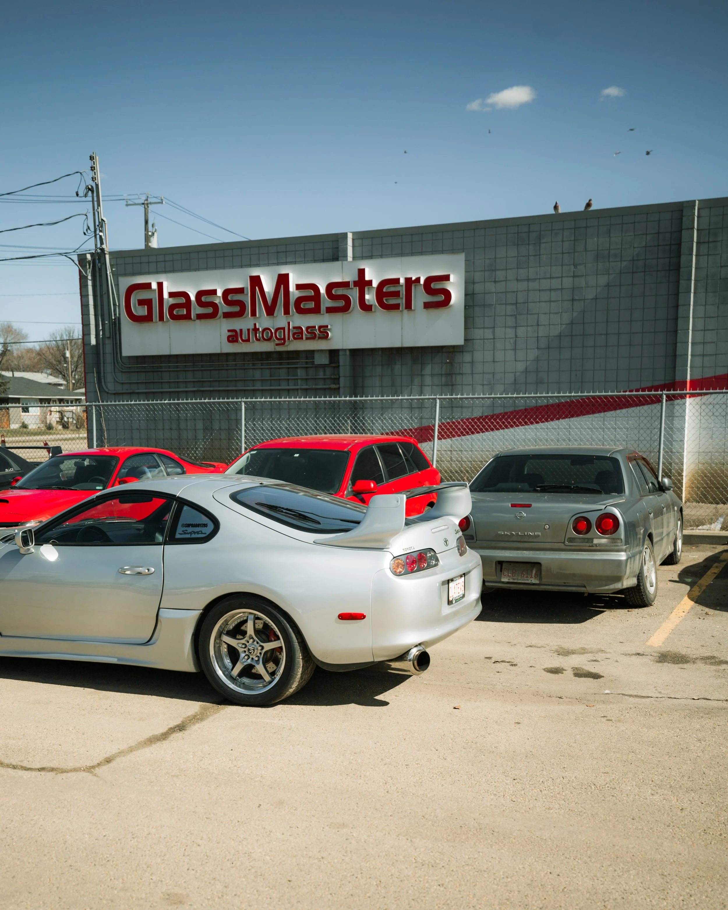 A parking lot with three cars parked in front of a building with a sign that reads 'GlassMasters auto glass' under a blue sky with some clouds and birds.
