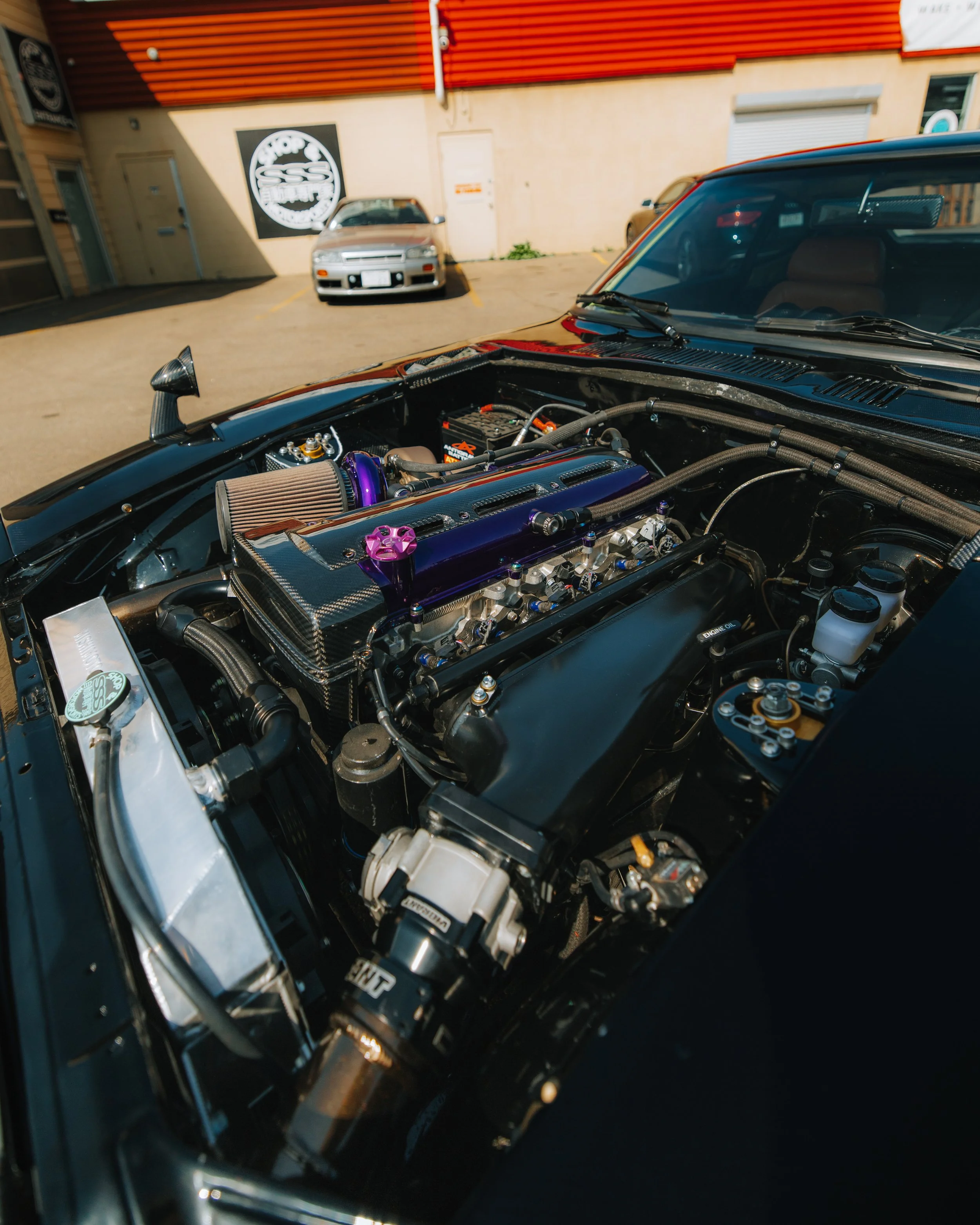 Close-up view of a modified car engine, featuring an aftermarket intake, carbon fiber components, and colorful engine parts, parked outside a building with cars in the background.