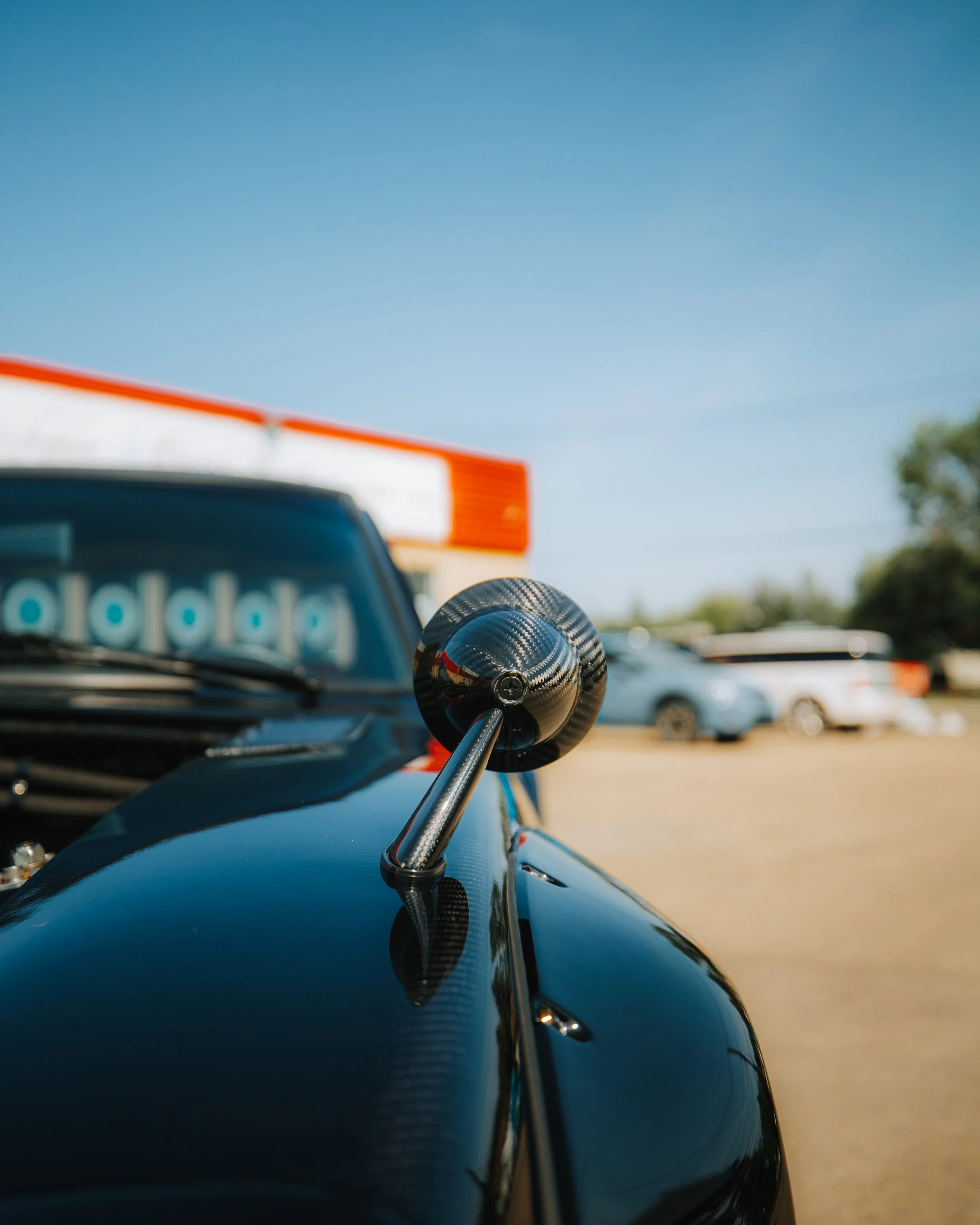 Close-up of a racing car's side mirror with a shiny, black, carbon fiber finish, reflecting the surrounding area, with other vehicles and a building in the background under a clear blue sky.