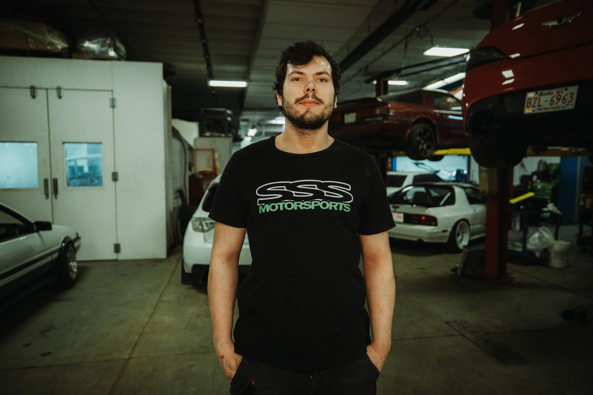 A man with dark, curly hair and a beard standing in a garage, wearing a black T-shirt with 'SSS MOTORSPORTS' printed on it. Cars are parked and lifted on racks in the background.