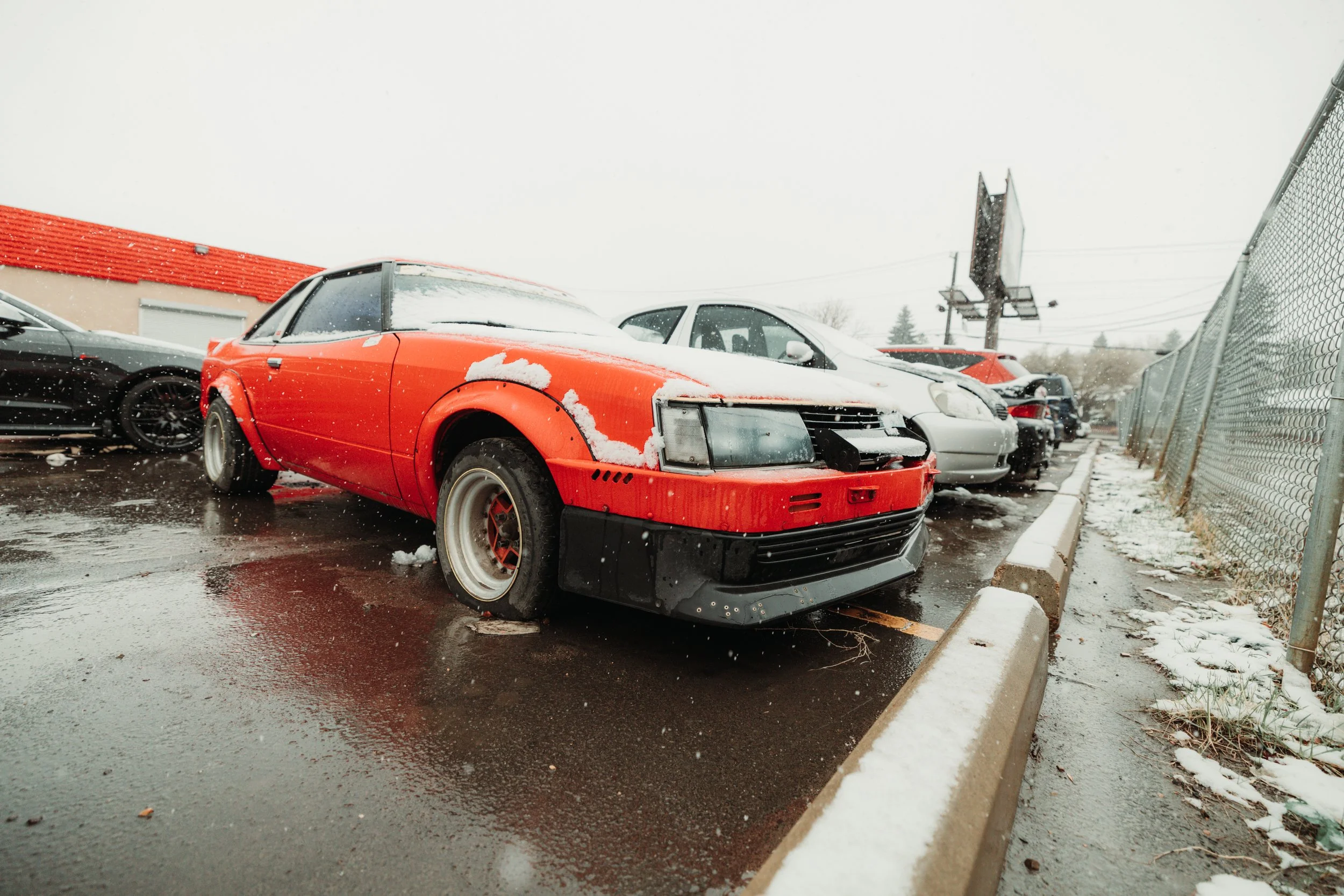 Red vintage sports car with snow on it, parked in a row of cars in a wet parking lot during snowy weather.