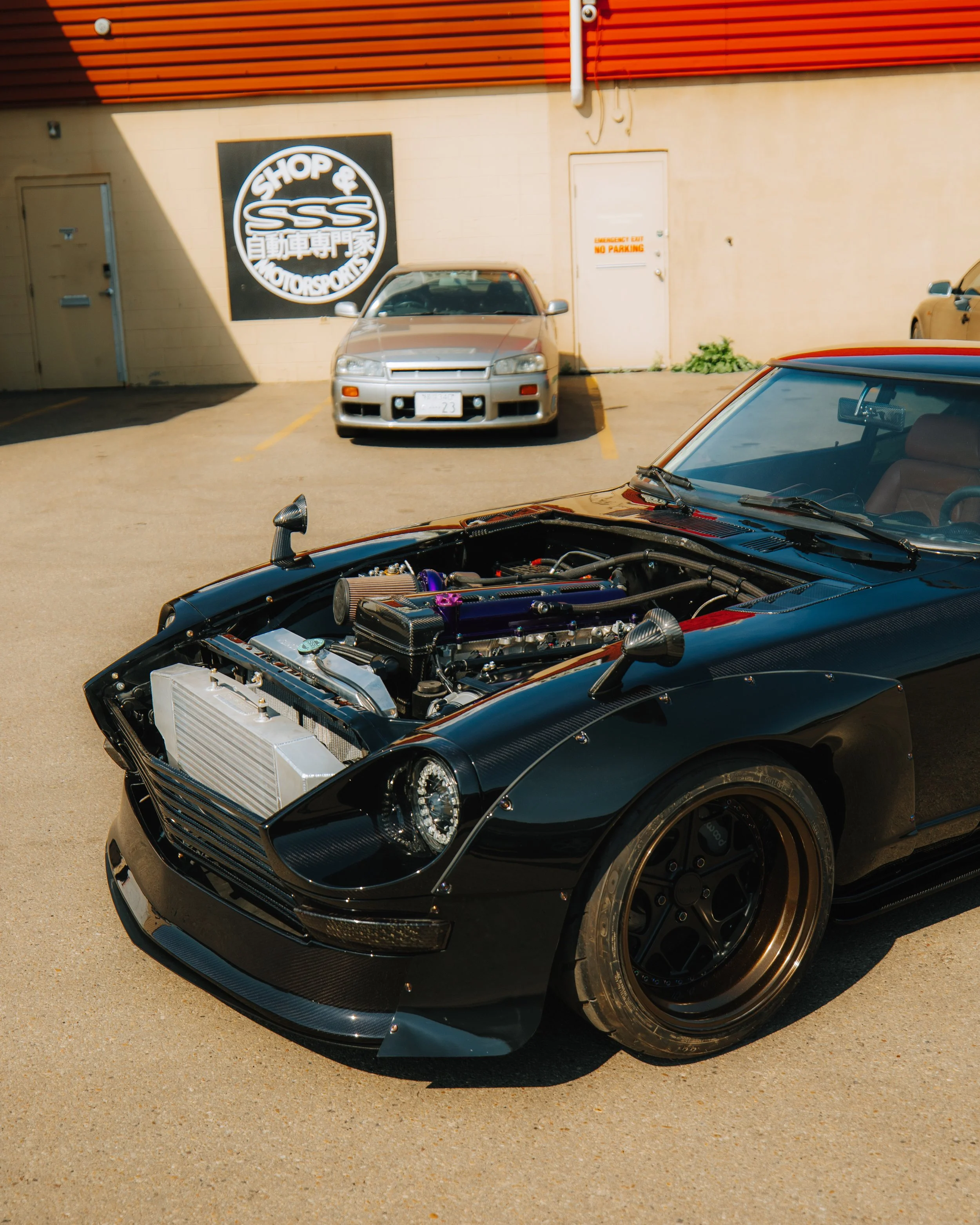 A black sports car with its hood open revealing a modified engine, parked in a lot with other cars and a building with a sign that says 'Shop & SSS Motorsports' behind it.