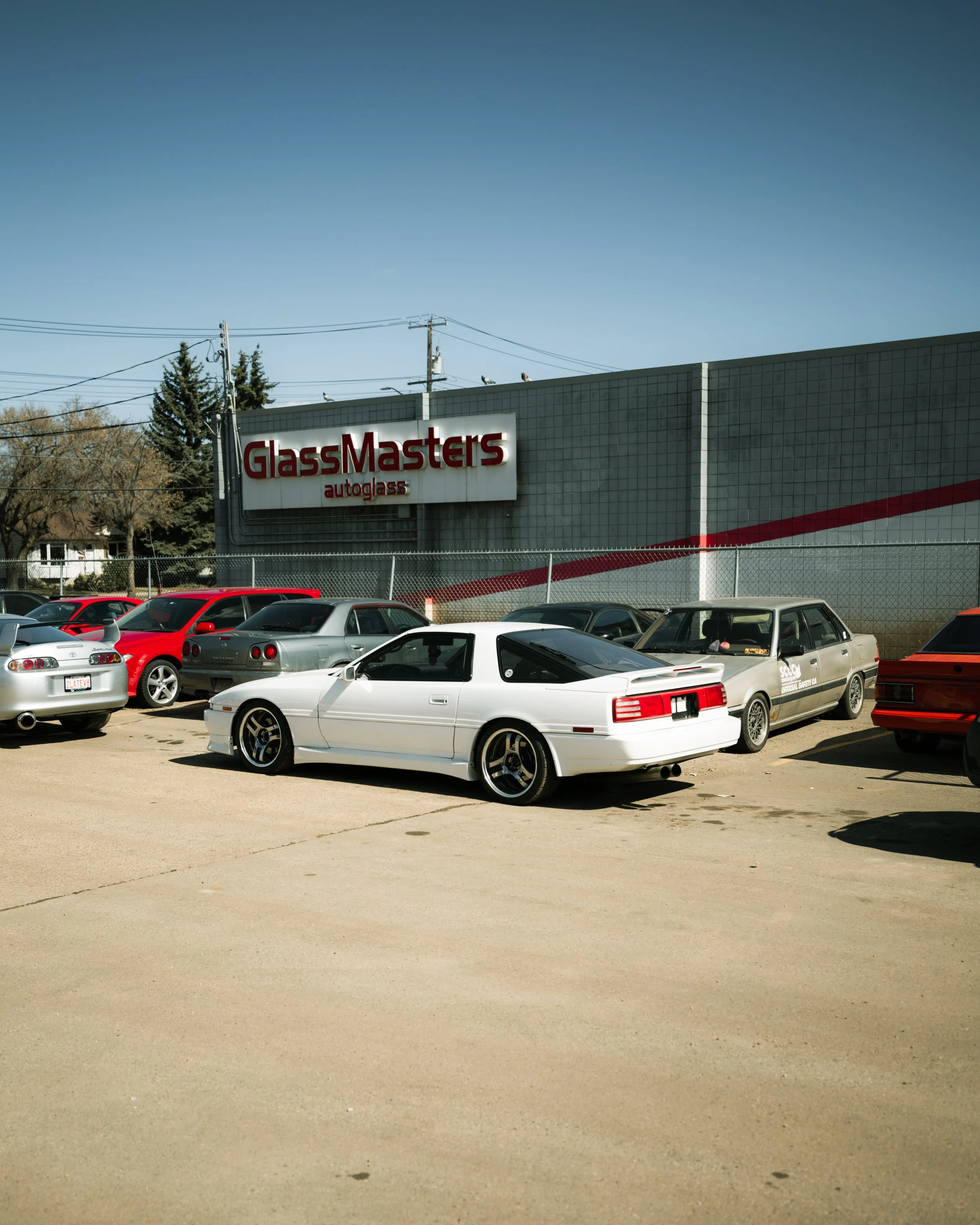 A parking lot in front of a GlassMasters auto glass shop with several cars, including a white sports car, parked on a sunny day.