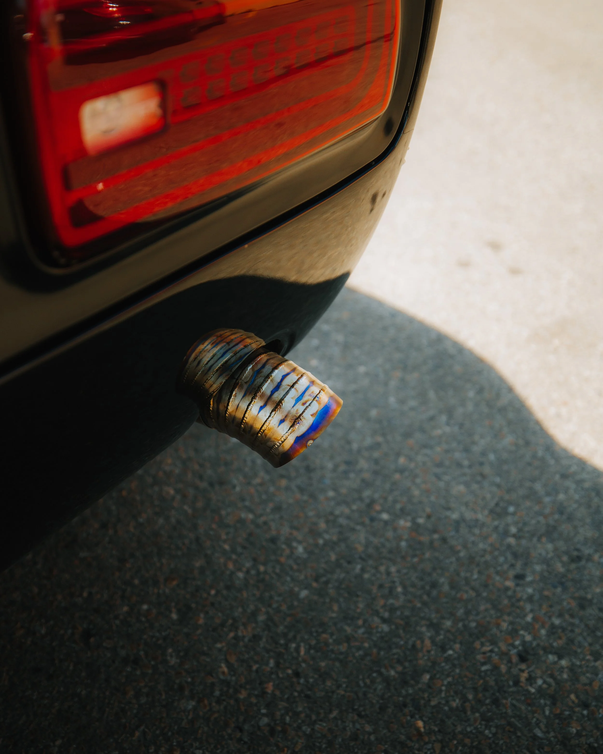 Close-up of a car's exhaust pipe with a burnished metal finish, under a rear tail light next to a textured asphalt surface.
