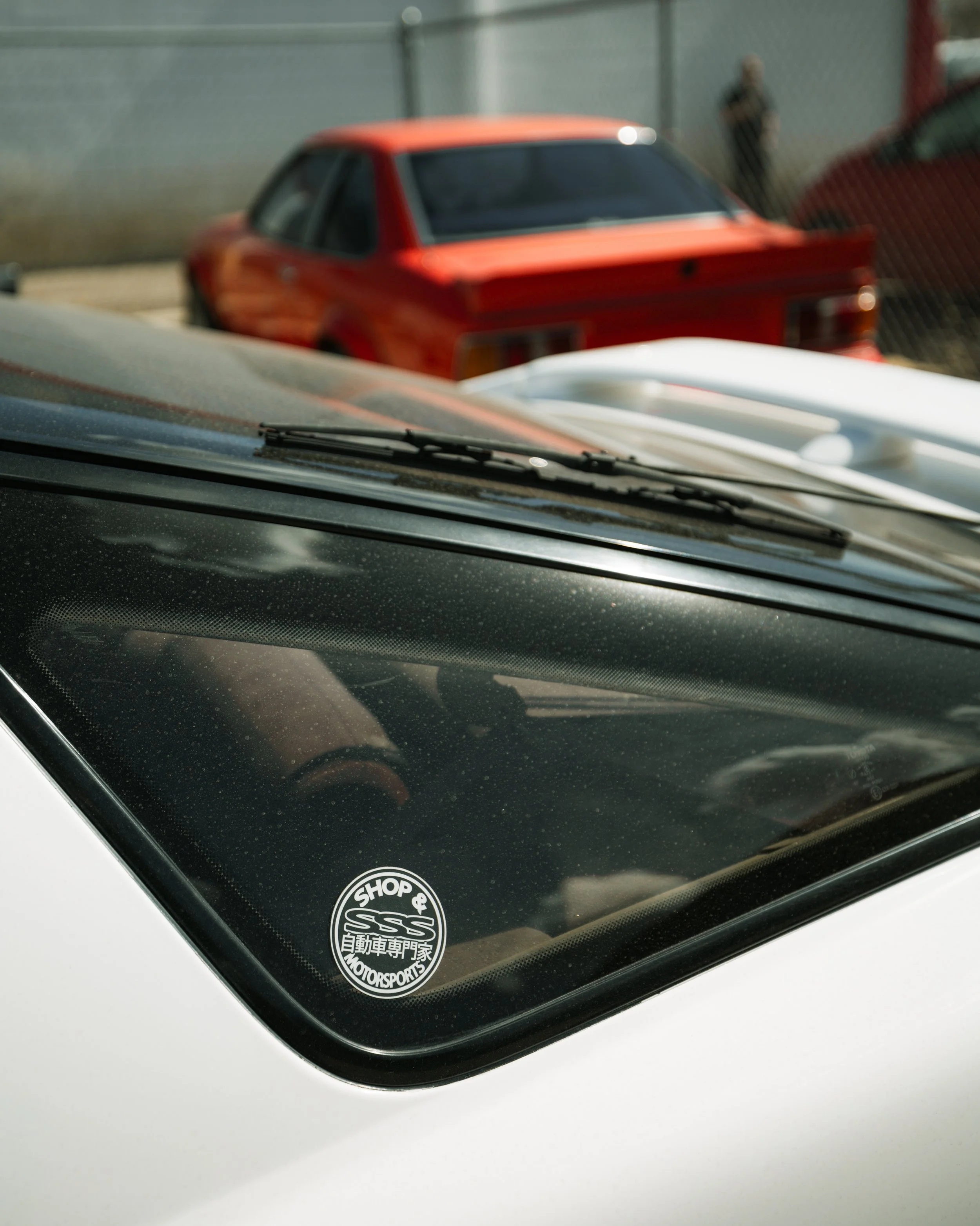 Close-up of a car's windshield with a sticker that says 'Shop & SSS Motorsports,' with three cars in the background, including a red and a white car.