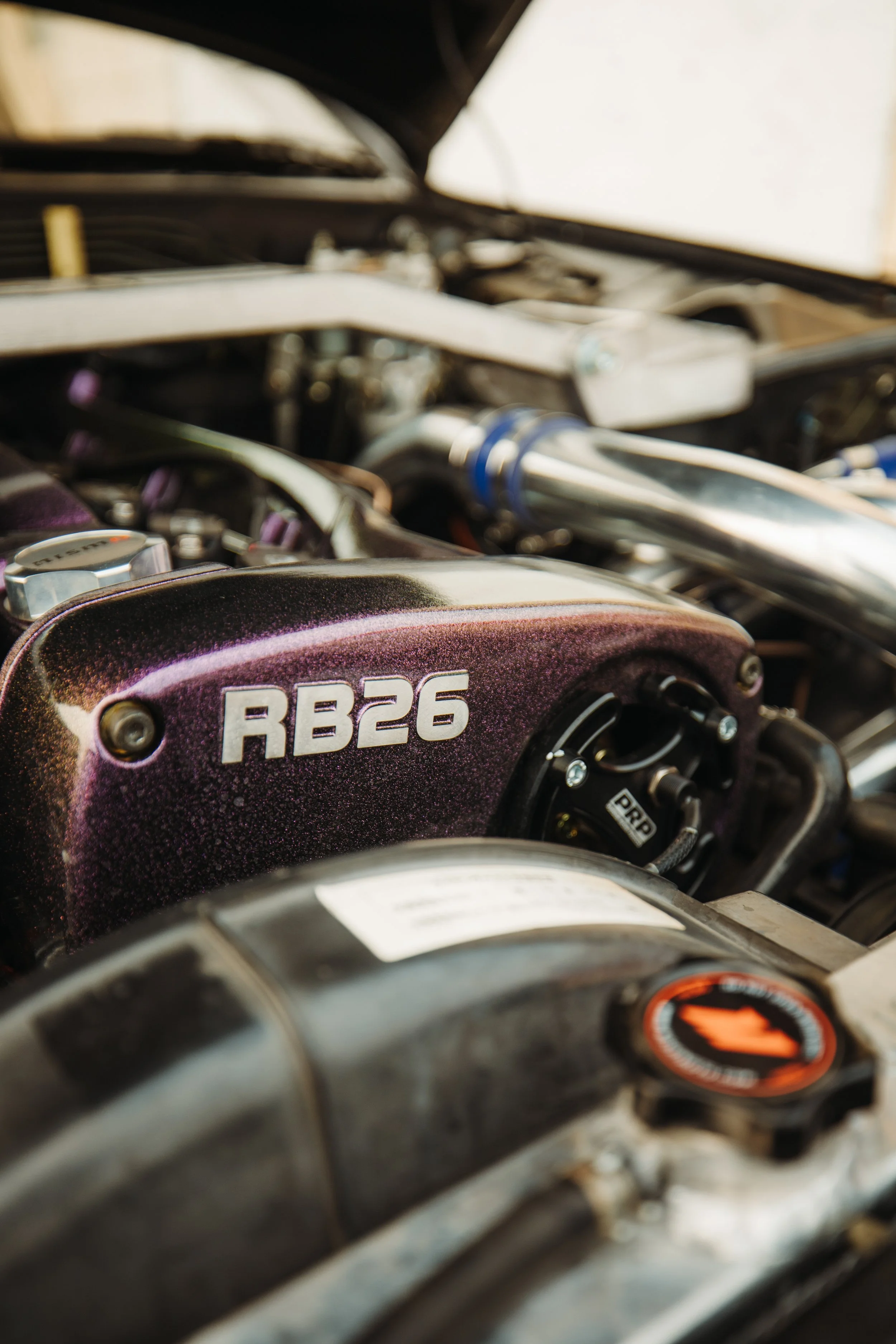 Close-up of a car engine with a pinkish-purple cover labeled RB26, indicating a Nissan RB26 engine.