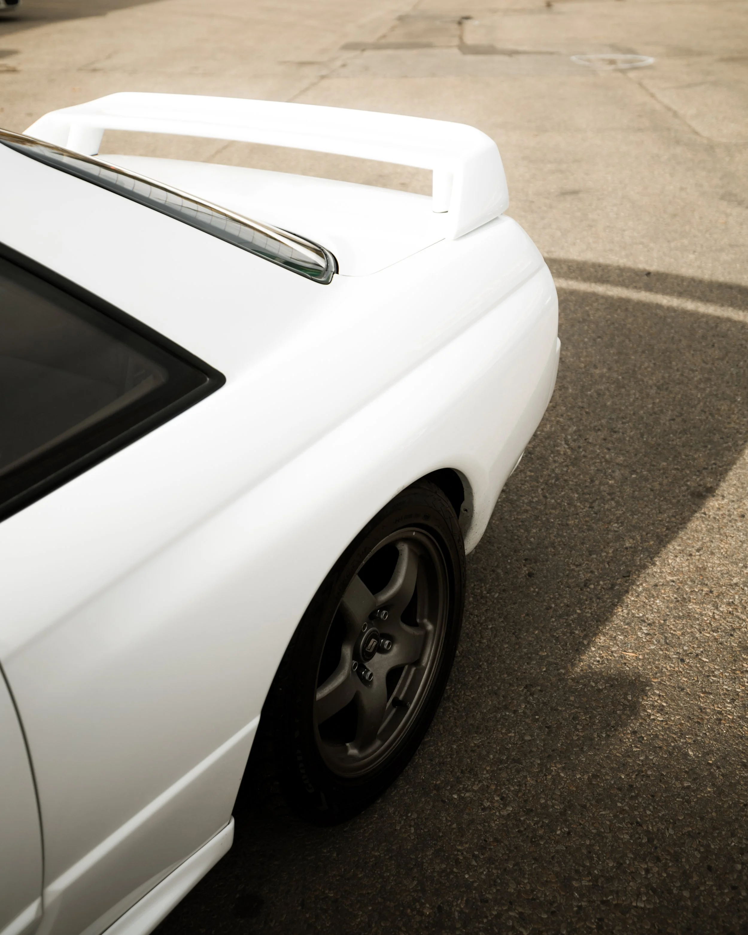 Close-up of the rear side of a white sports car, showing the rear wheel, spoiler, and part of the tinted window, parked on a street.