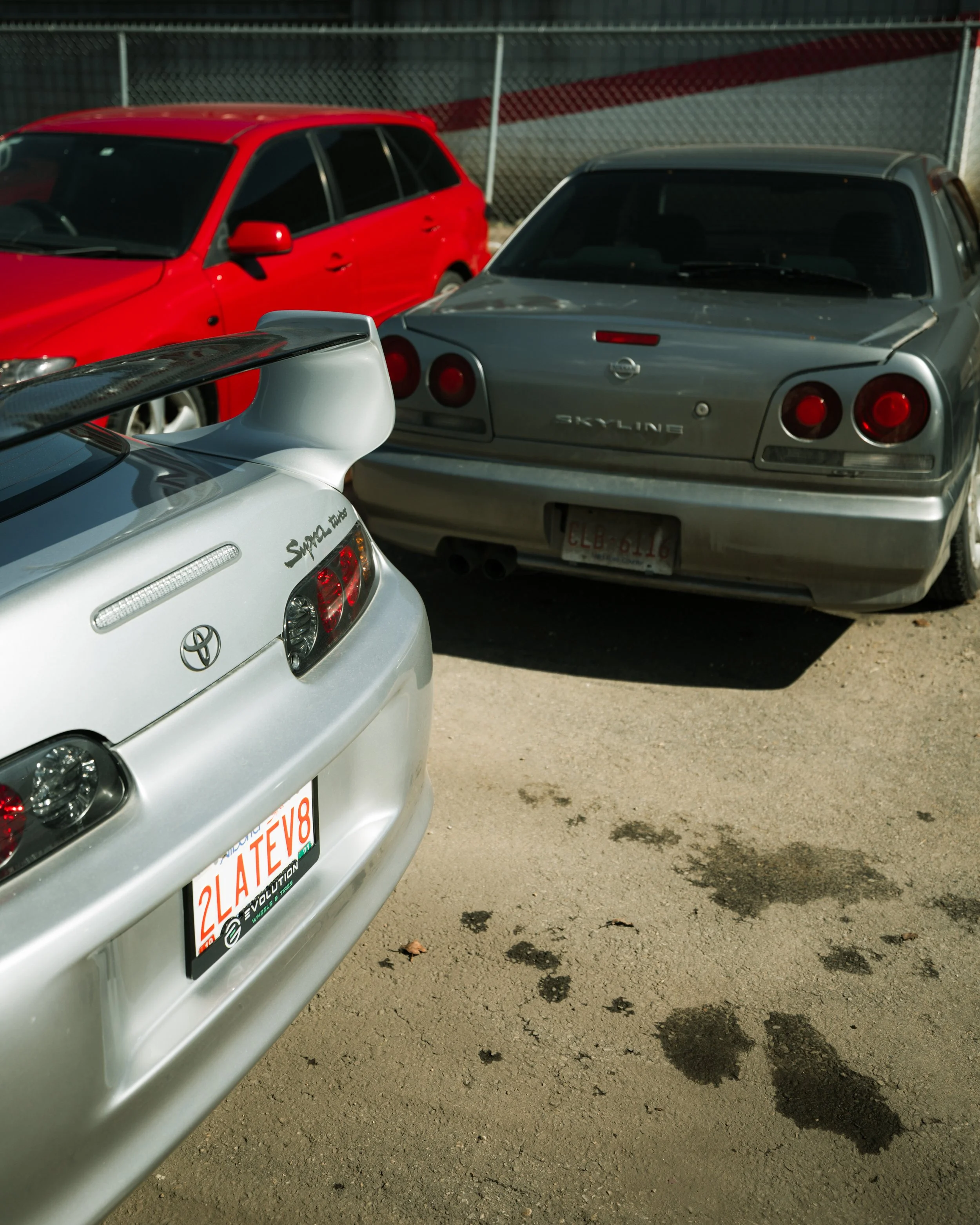 Three parked cars, including a silver Toyota Supra with a large rear wing, a gray Nissan Skyline, and a red station wagon, in a lot with a chain-link fence and dirt ground.