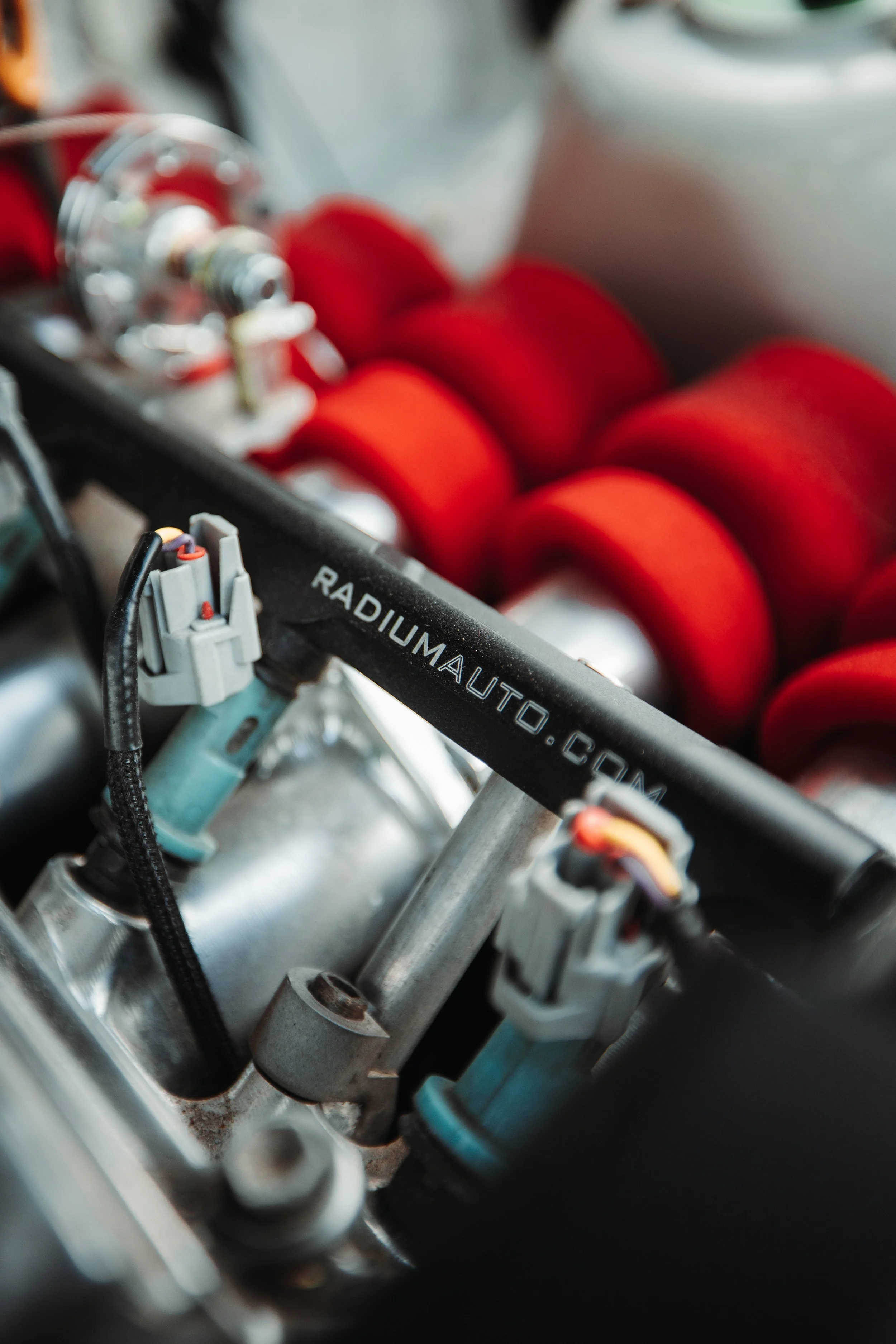 Close-up of a racing engine with red components and a black metal rod labeled 'RADIUMAUTO.COM' connected to various sensors and parts.