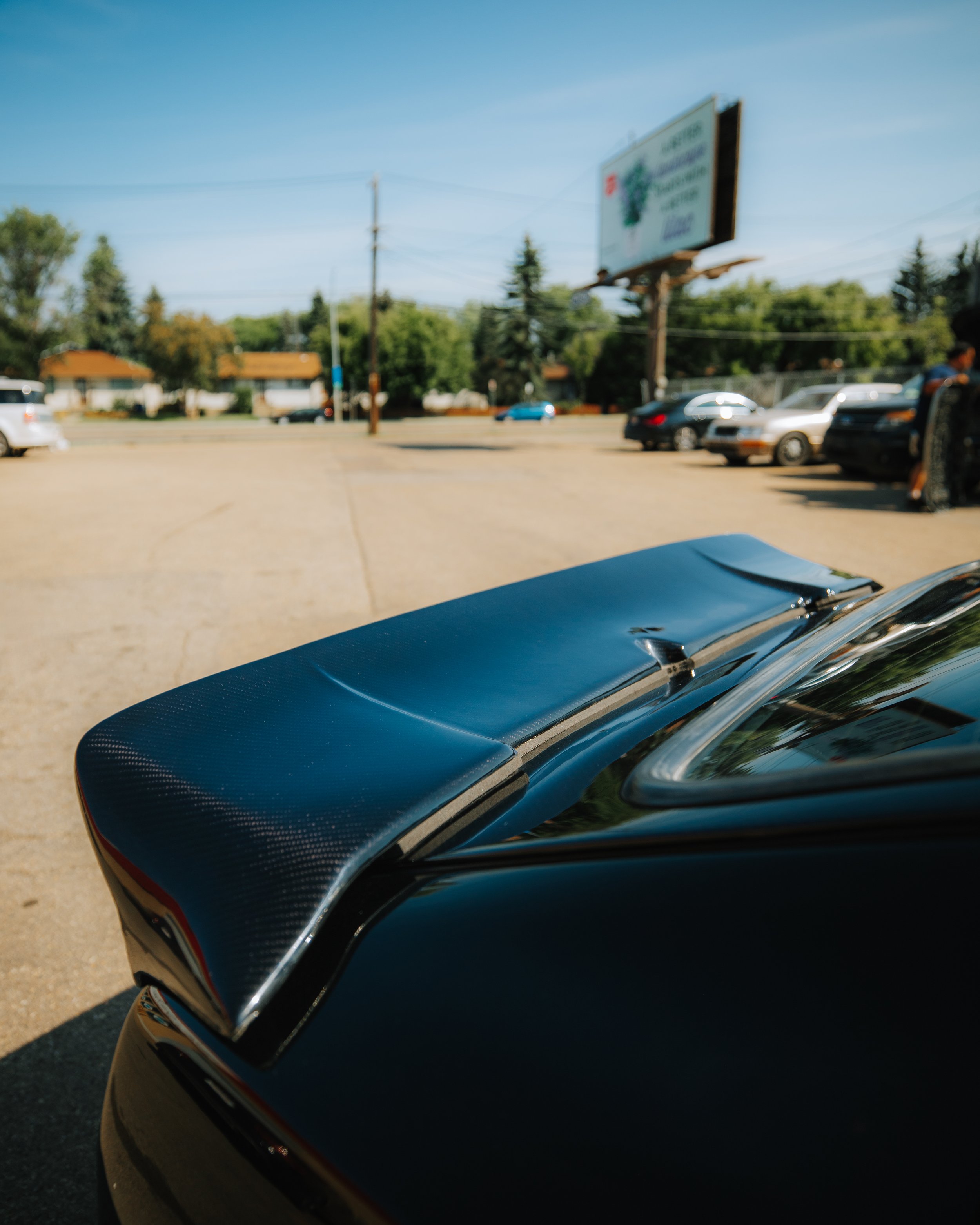 Close-up of a black car's rear spoiler with a carbon fiber texture in a parking lot with other cars and a billboard in the background.