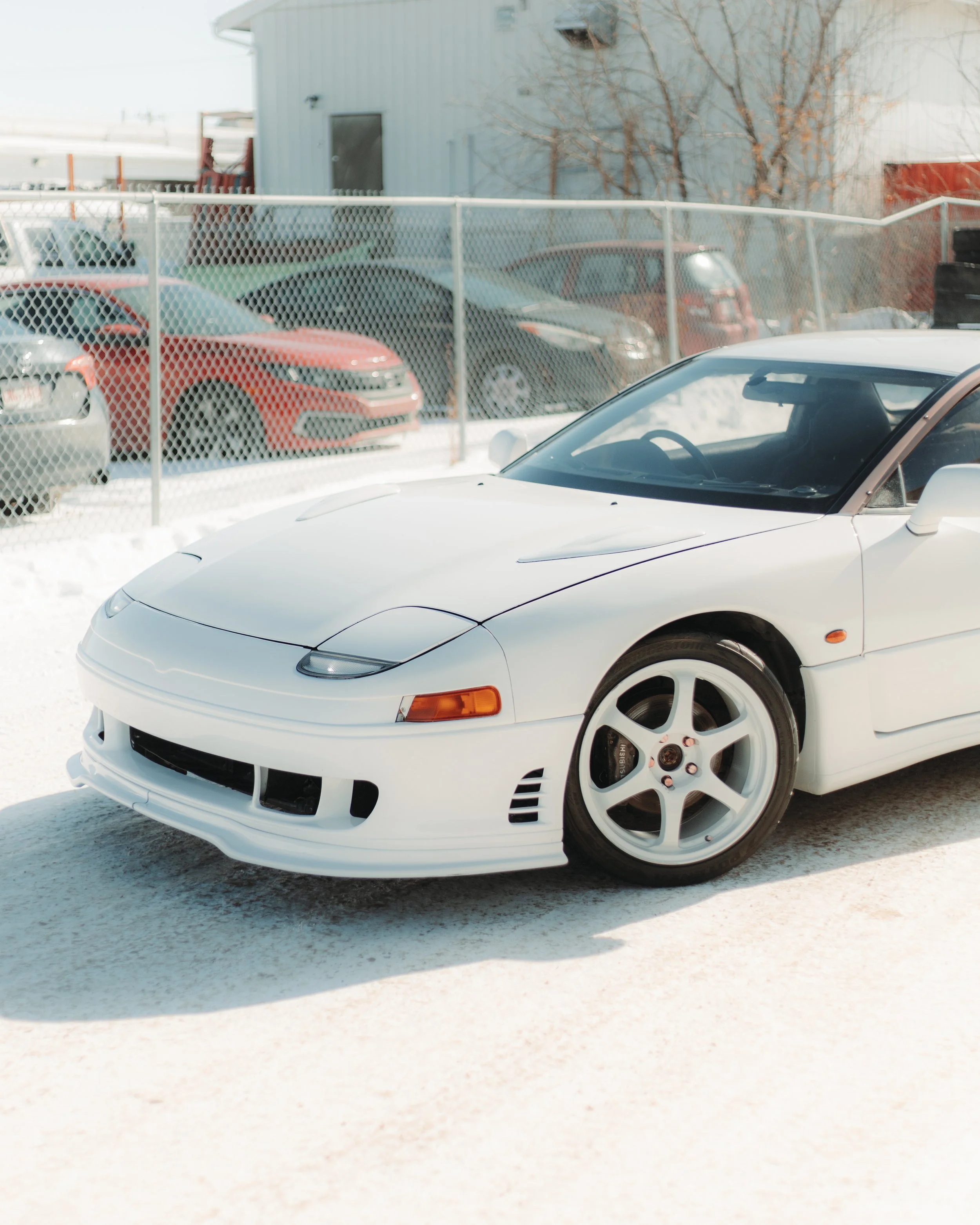White sports car parked on snow with a chain-link fence and other cars in the background.