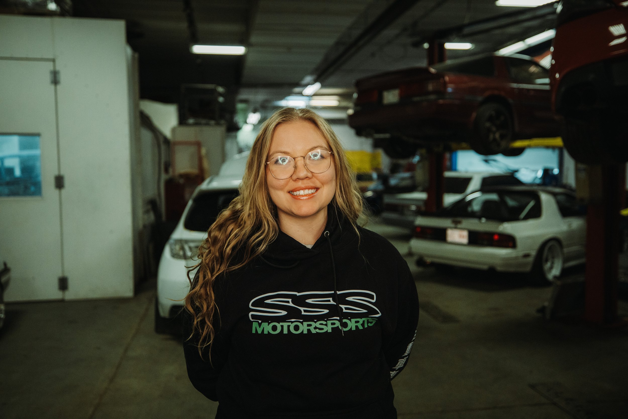 A smiling woman with long curly hair and glasses standing in a car repair garage, with various cars and car lifts in the background.