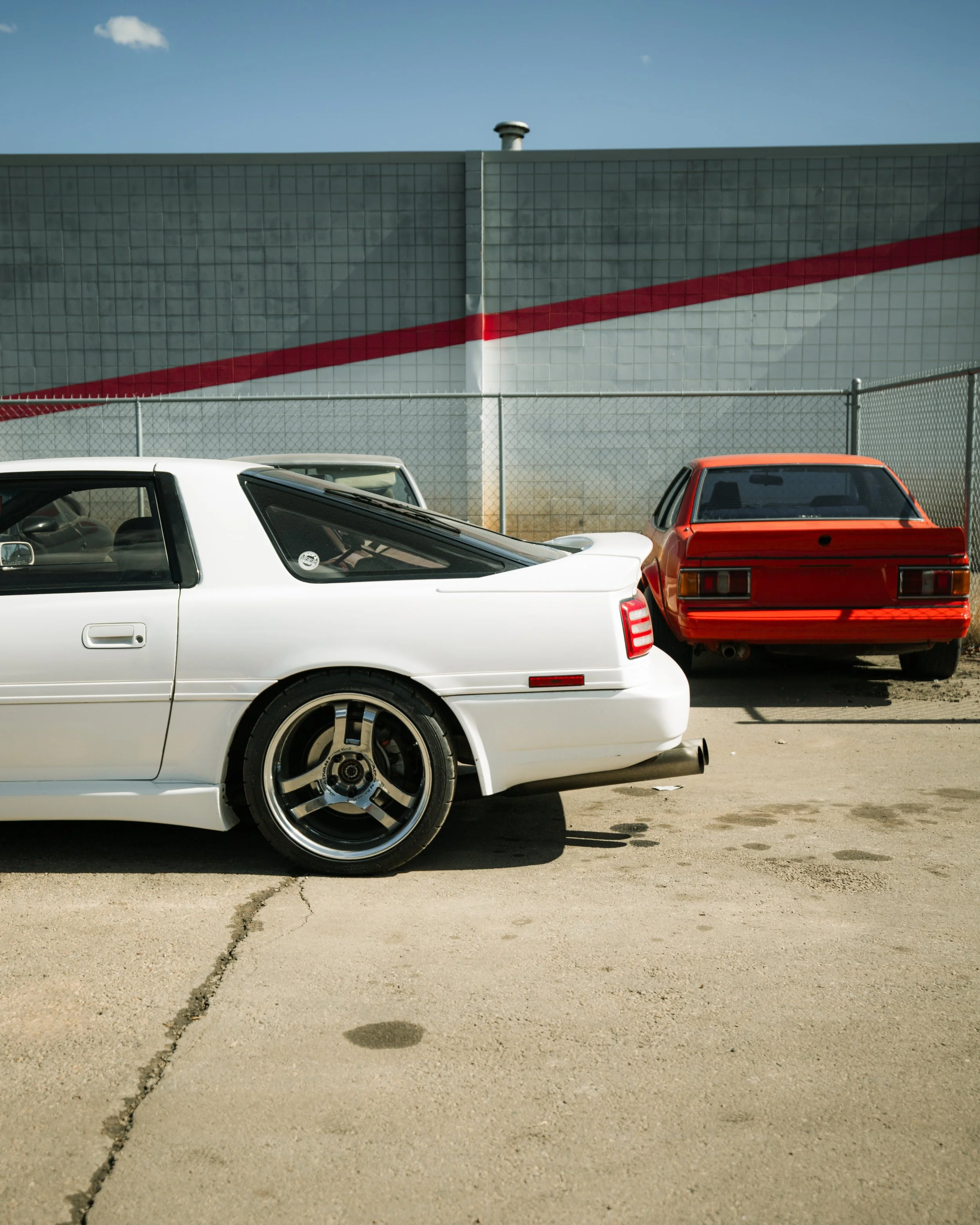Two parked cars, a white sports car in the foreground and a red sedan behind it, in front of a chain-link fence and a gray industrial building with a red stripe.