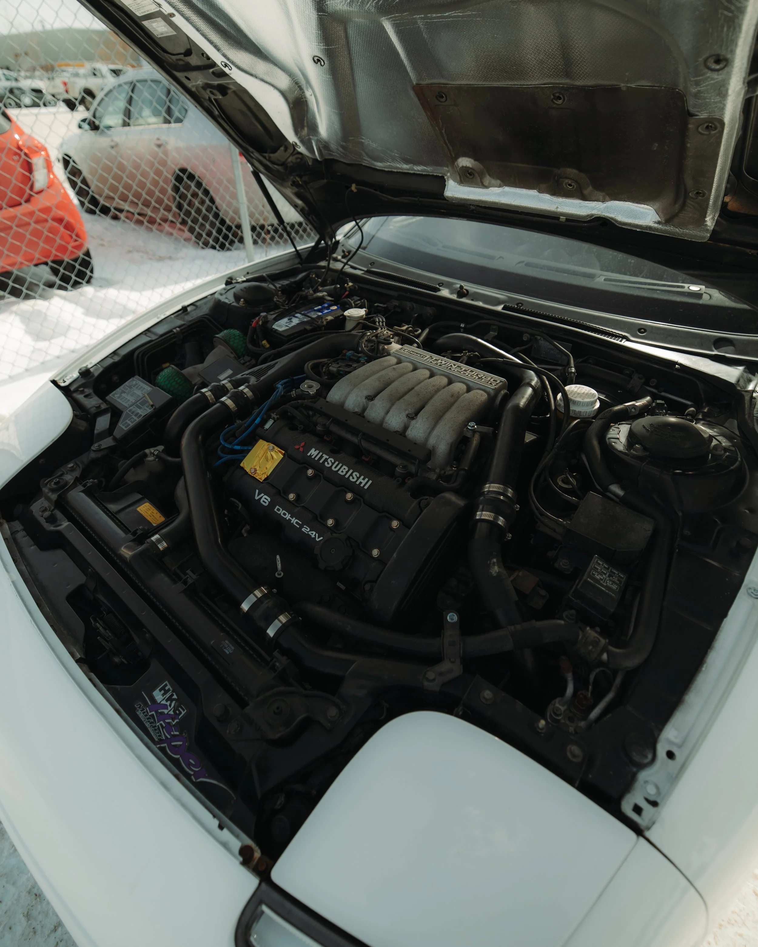 Open hood of a white Mitsubishi sports car revealing the engine bay, with visible engine components and hoses.