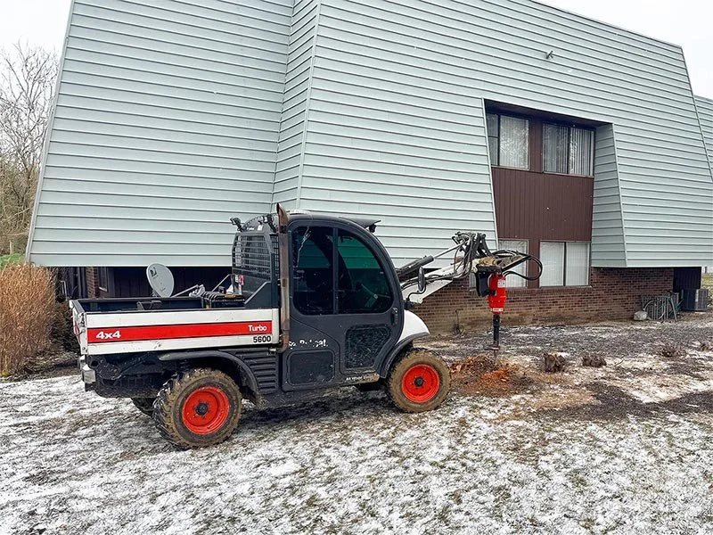 A small Bobcat compact utility loader with a hydraulic auger attachment drilling into the ground outside a building with a gray facade, some snow on the ground, and trees in the background.