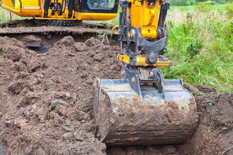Close-up of a yellow excavator bucket digging into the dirt at a construction site with green grass in the background.