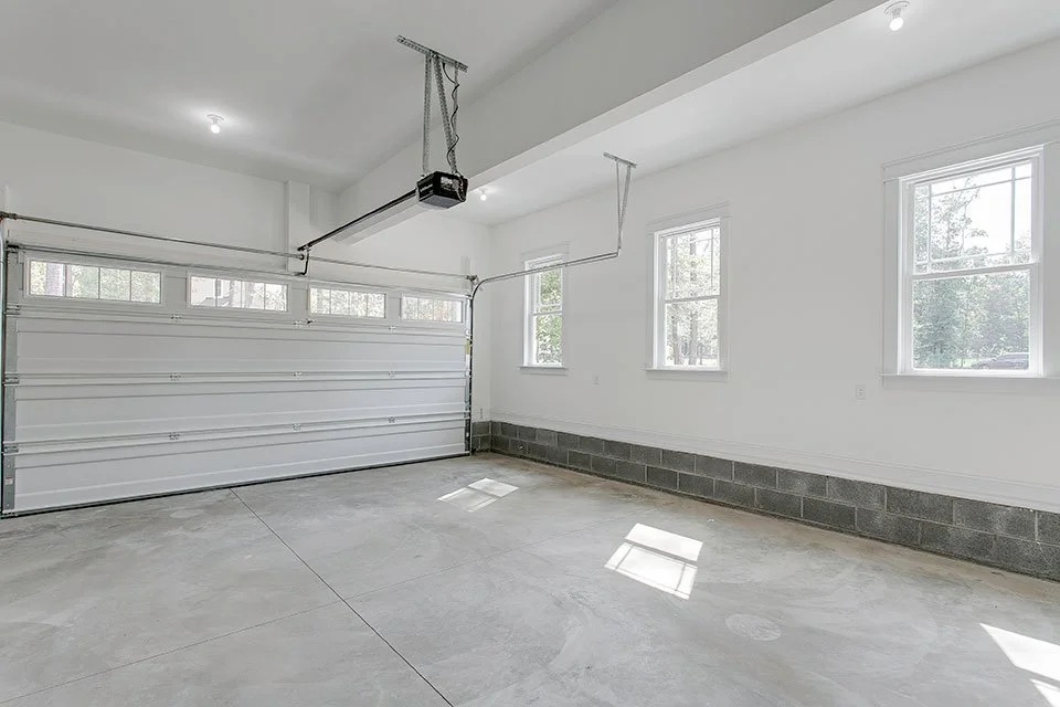 Empty garage with white walls, concrete floor, and a white roll-up garage door. Several windows allow natural light inside.