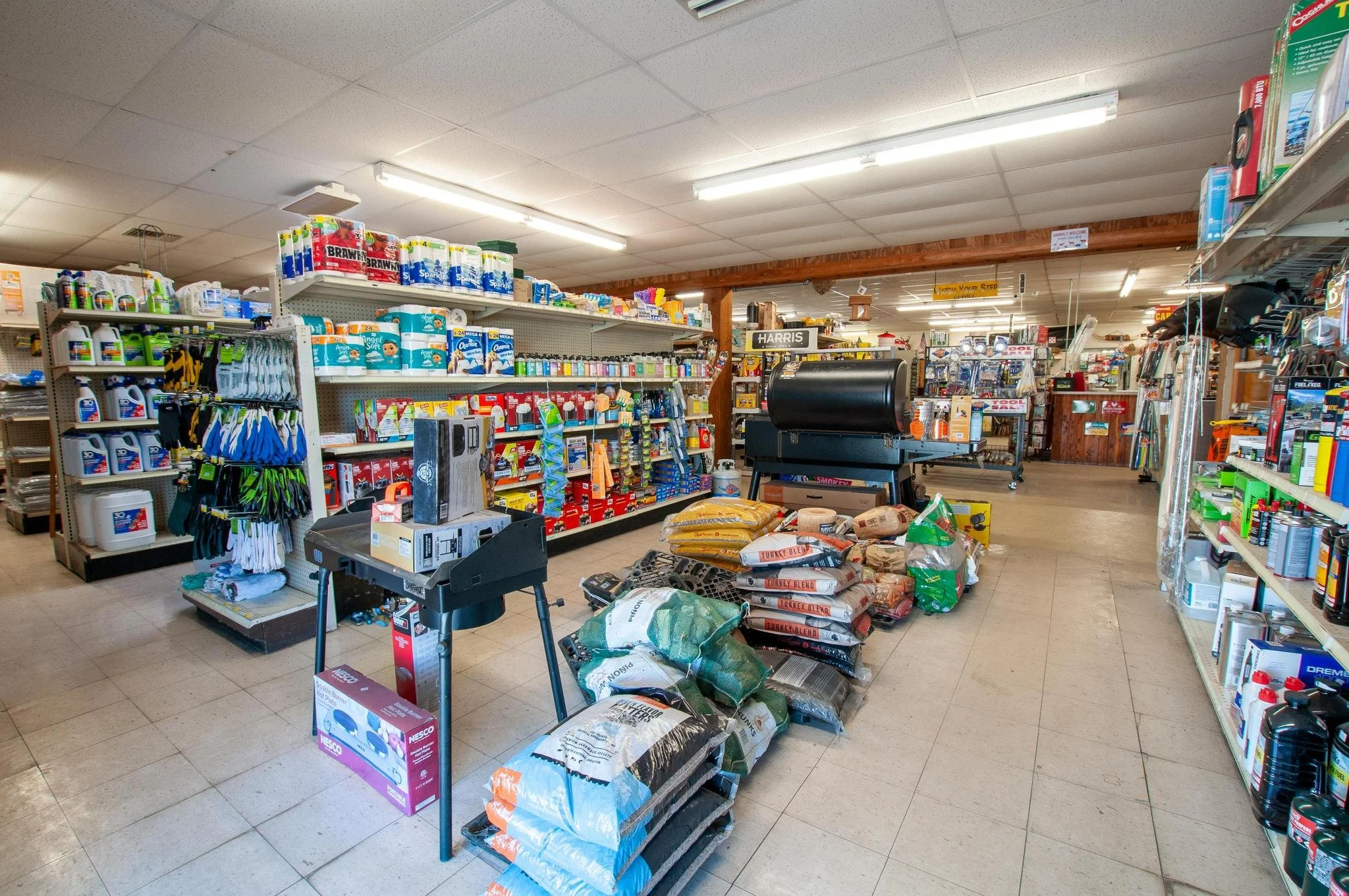 Interior of a hardware store with shelves stocked with various garden and household supplies, bags of soil and fertilizer on the floor, and gardening tools visible.