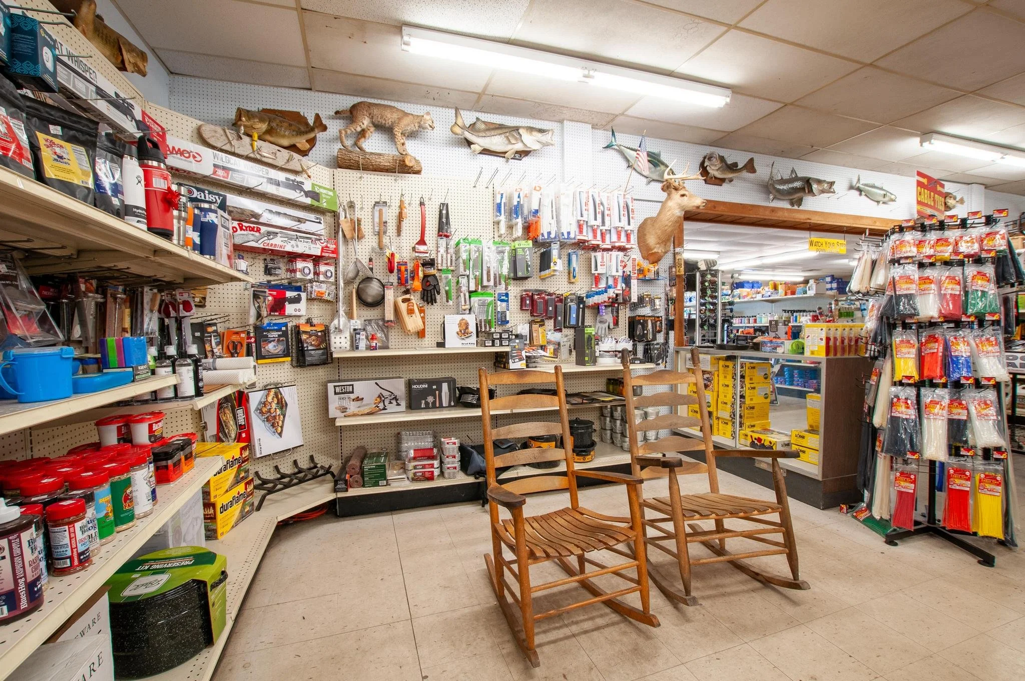 A hardware store aisle with hunting and fishing supplies, including mounted animal heads, fishing lures, knives, and tools. Two wooden rocking chairs are in the foreground.