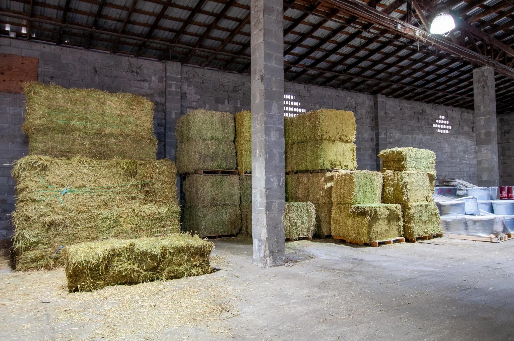 Stacks of hay bales stored inside a barn with a concrete pillar in the center, a corrugated metal roof, and a concrete floor.