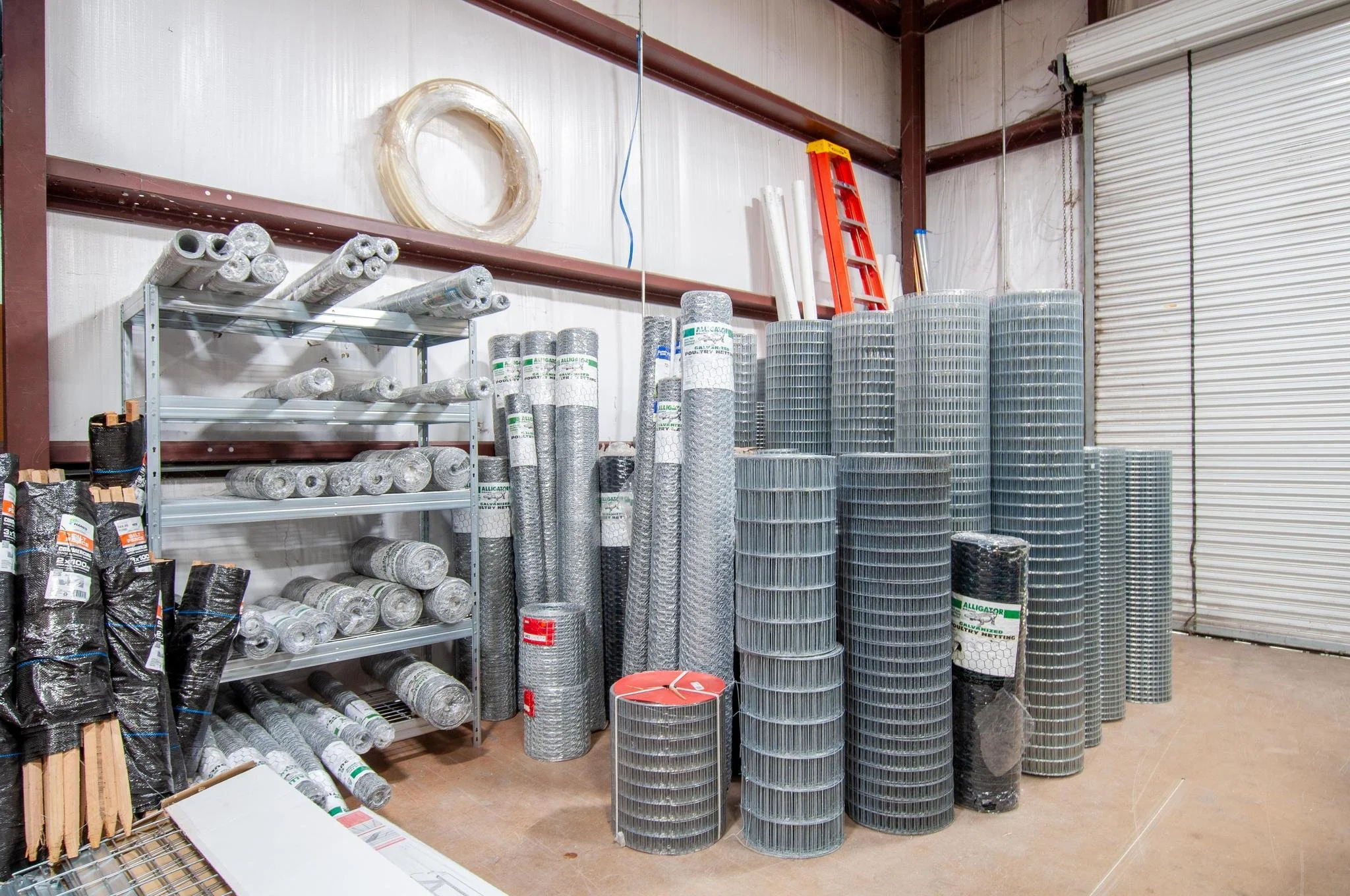 Storage room with rolled wire fencing panels and other fencing materials, including rolls of wire mesh, stacked against the wall on an industrial shelving unit, with a red ladder leaning against the wall and a roll of white tubing on the shelf.