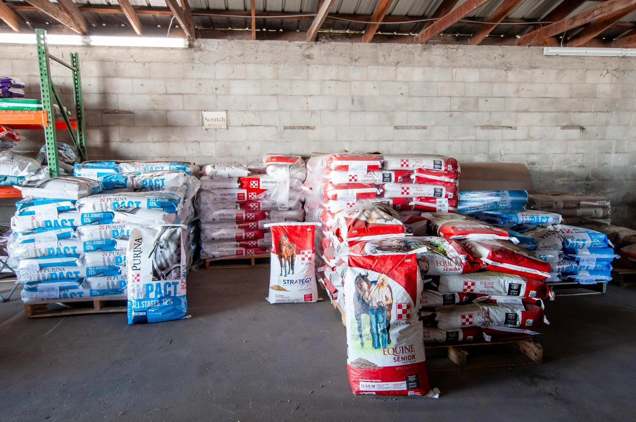Shelves of large bags of dog and horse feed stacked in a warehouse with a brick wall and wooden ceiling.