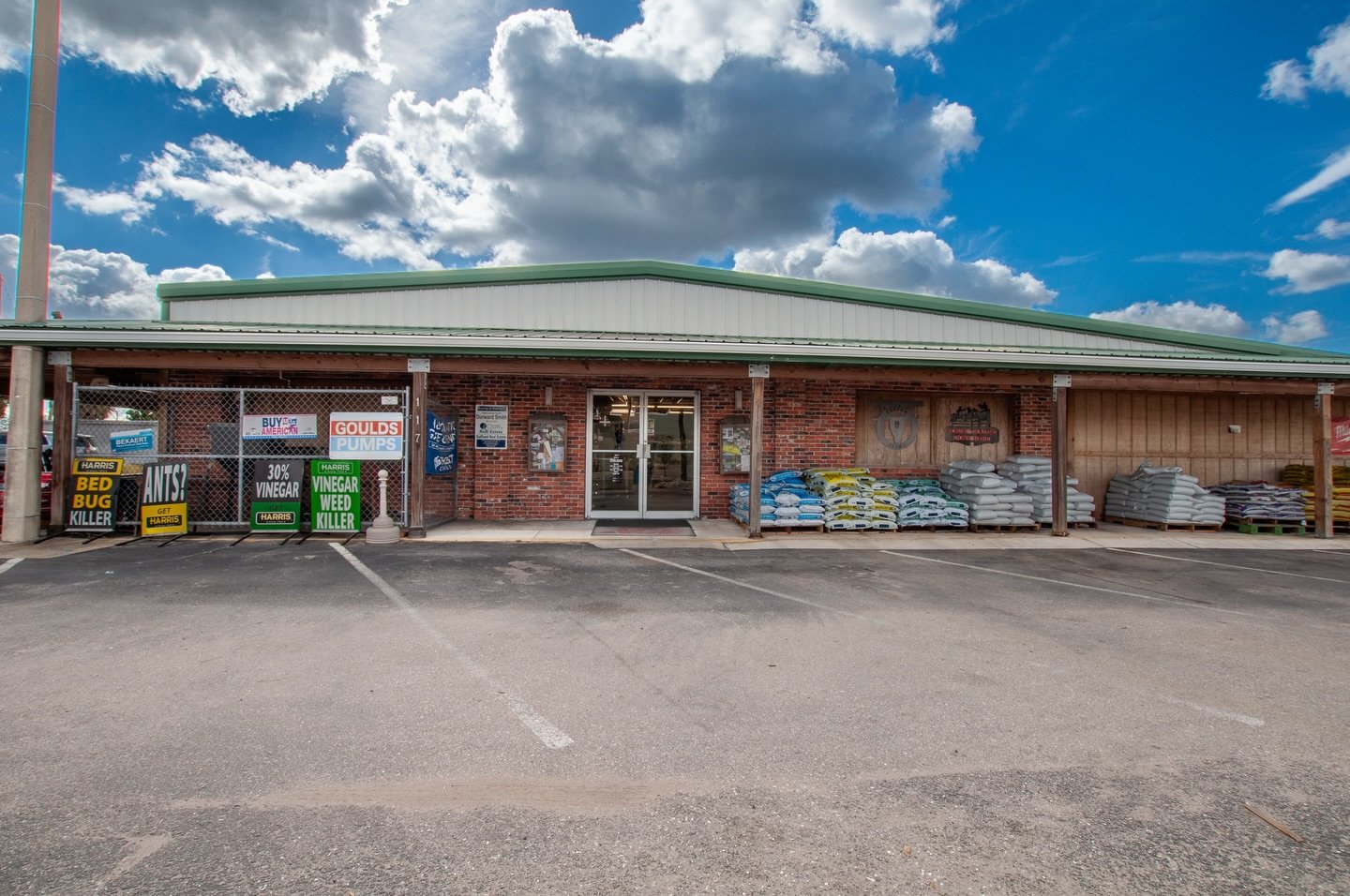 Exterior of a gardening store with bags of soil or fertilizer stacked outside, signage advertising pest control and weed killer, parking lot in the foreground, partly cloudy sky overhead.