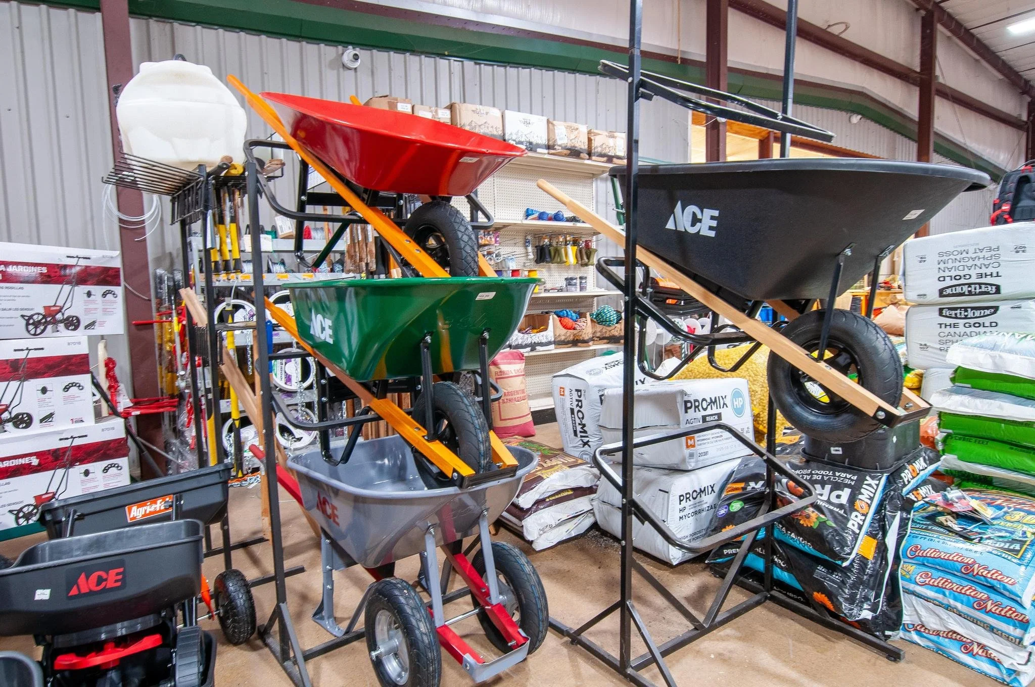 Display of various wheelbarrows including red, green, gray, and black, stacked and positioned in a hardware store, with shelves of gardening supplies and fertilizer bags in the background.