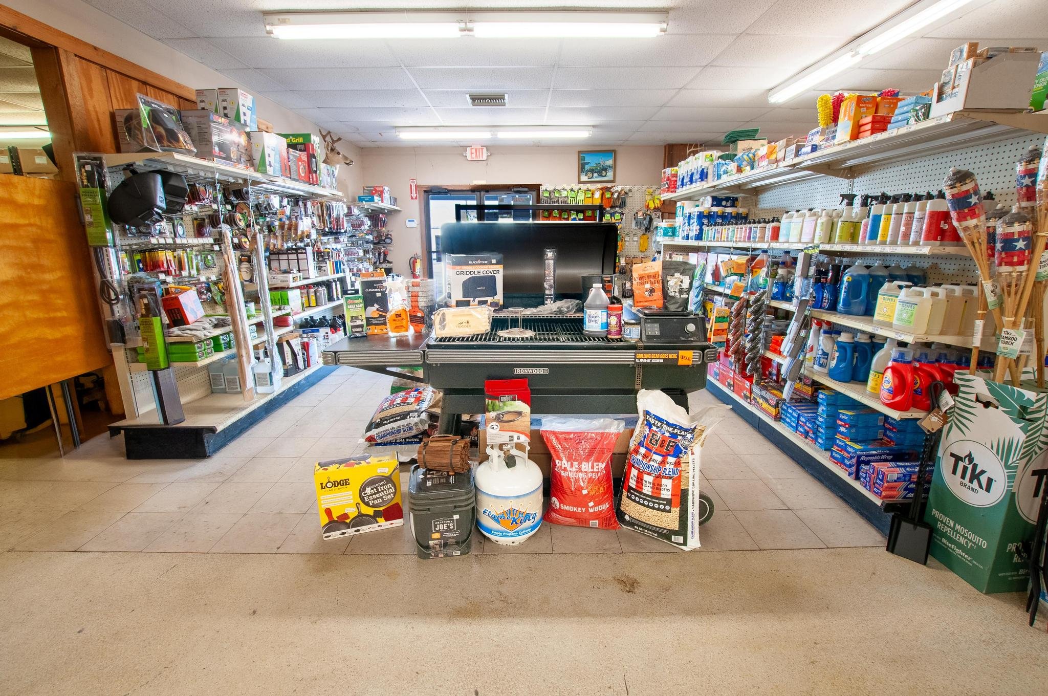 Store interior with shelves of various products, a grill on display in the center, and items like propane tank, bags of charcoal, and grilling accessories.