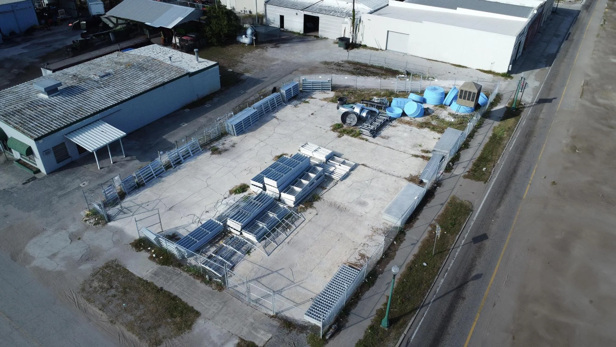 A fenced storage yard with stacked metal frames, blue water tanks, and industrial equipment, adjacent to a building, on a cracked concrete surface beside a road.