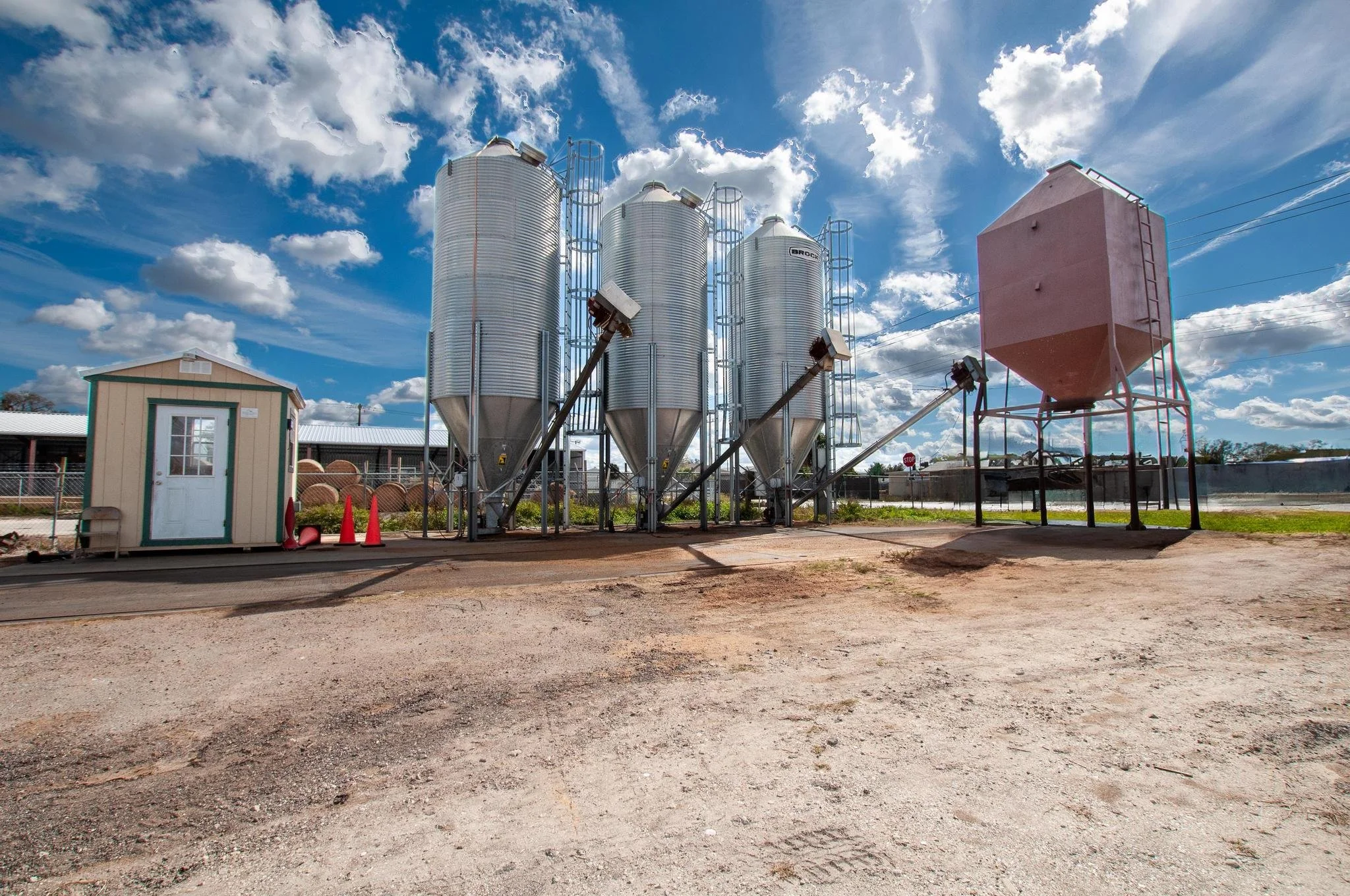 A farm or industrial setting with four large silver grain silos and a smaller pink grain bin on stilts, under a partly cloudy blue sky.