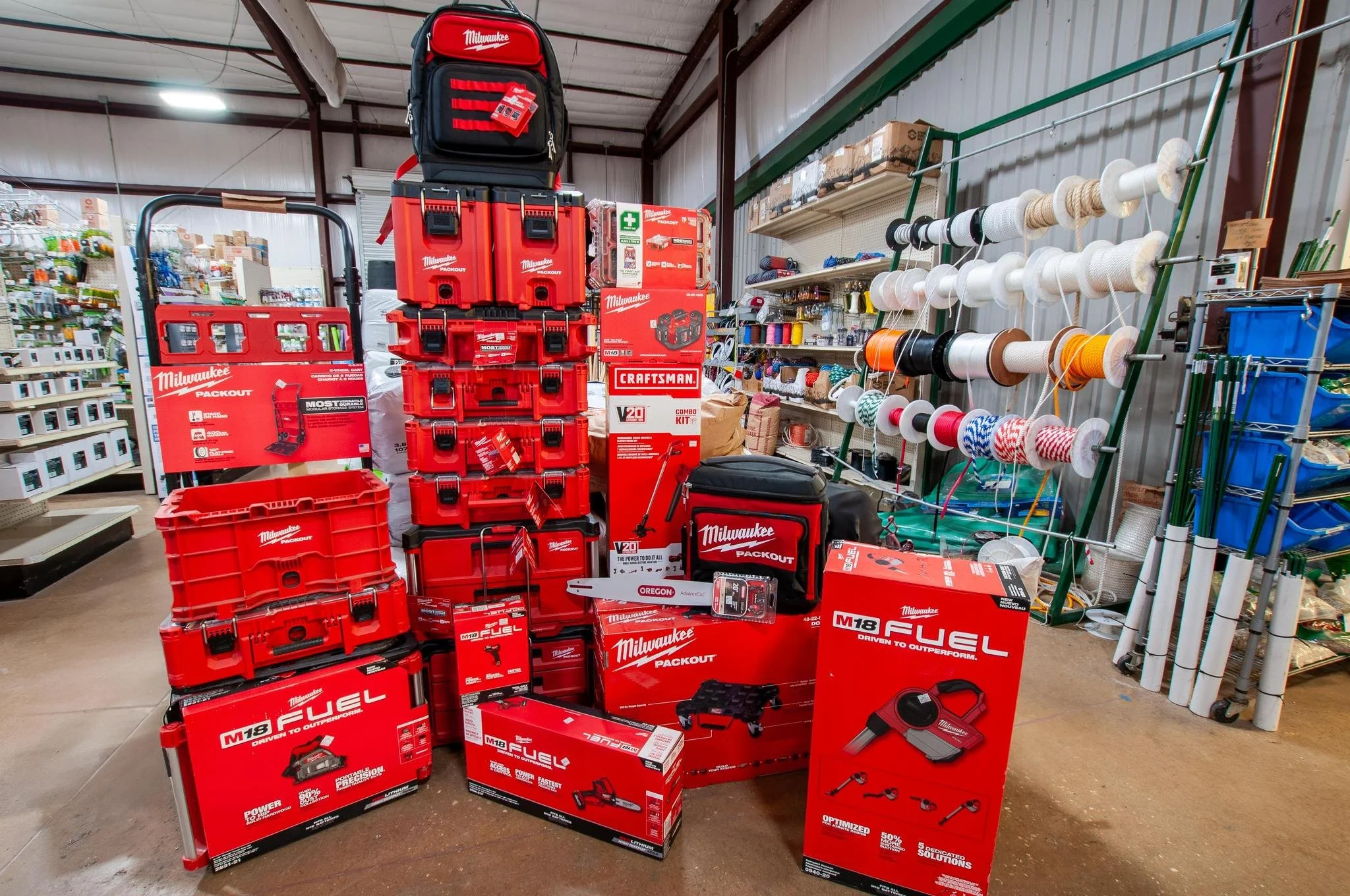 Display of Milwaukee power tools and accessories at a hardware store, including tool boxes, battery packs, and various drill sets, with rolls of rope and other supplies in the background.