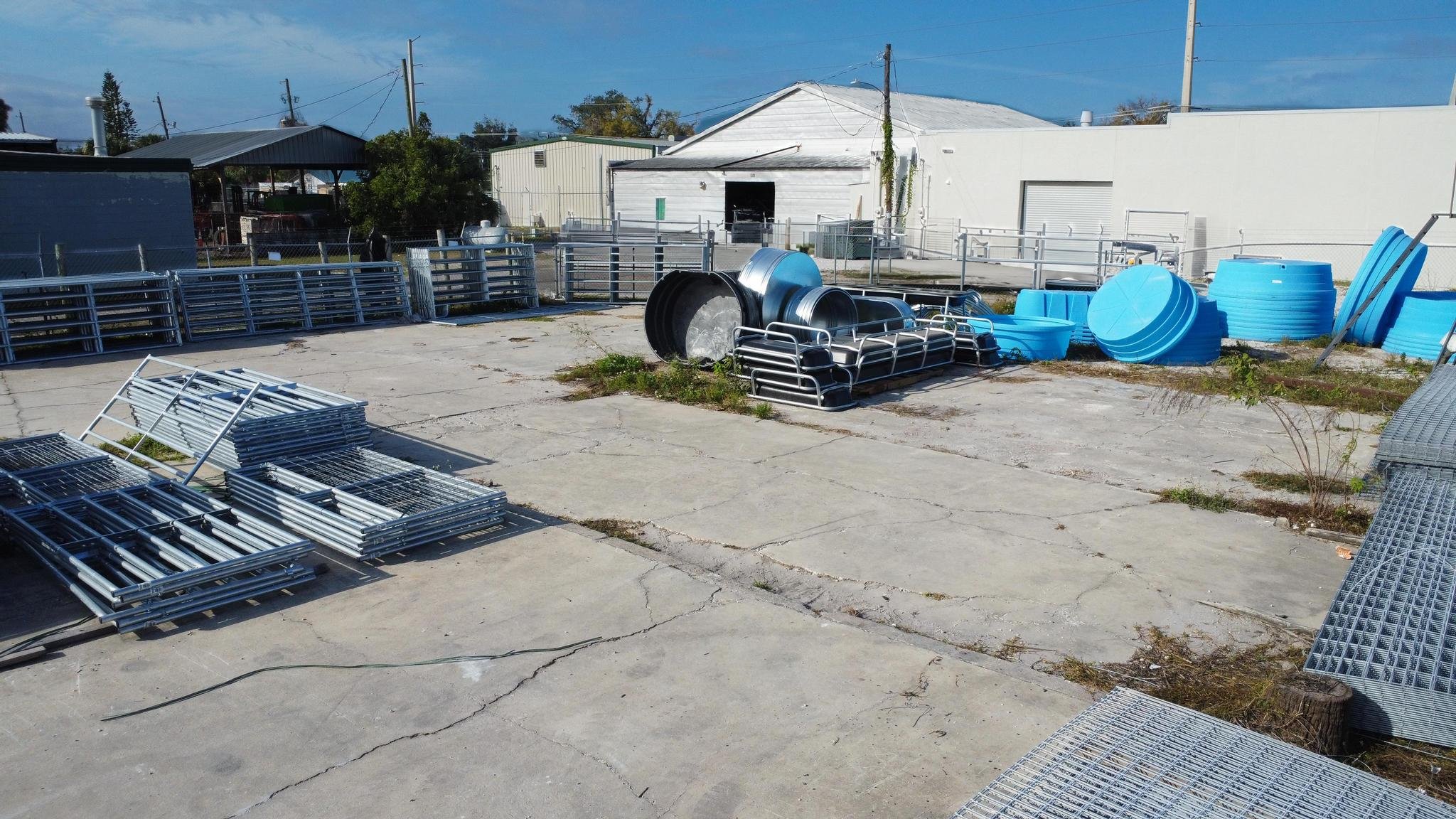 Outdoor storage yard with metal fencing, stacked blue plastic tanks, metal racks, and concrete pavement with cracks.