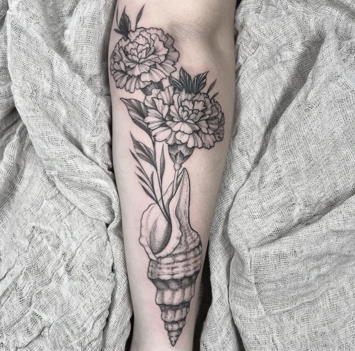 Tatuaje de dos flores de peonía con hojas en el antebrazo, con detalles en negro y gris, en un fondo de tela de color claro.