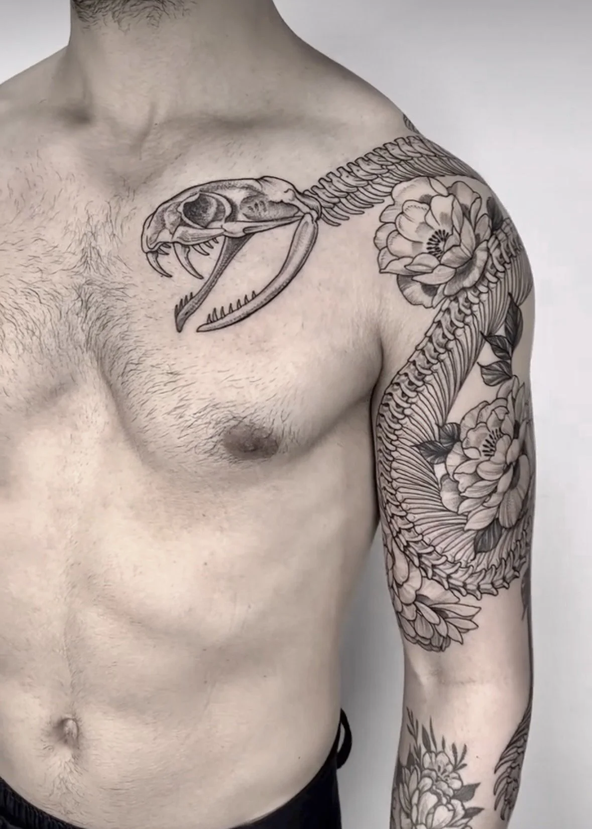 Tatuaje de un esqueleto de criatura prehistórica en el pecho y brazo de un hombre, con detalles en negro y gris, incluyendo flores en el brazo.