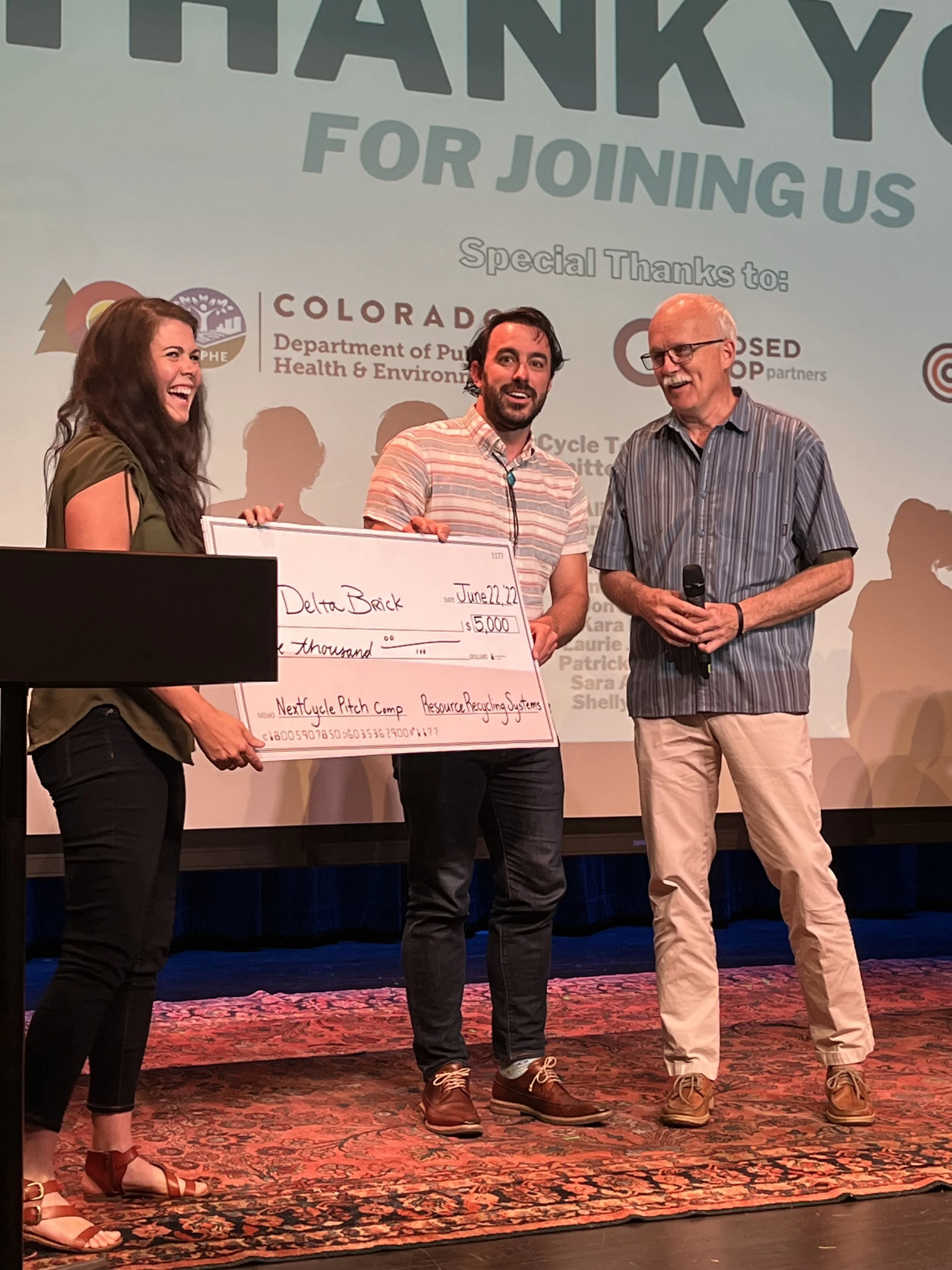 Three people stand on a stage at the NextCycle pitch competition. A woman on the left and the man in the middle are holding an oversized grand-prize check. An older man on the right holds a microphone.
