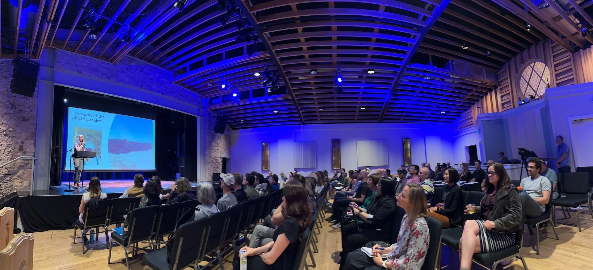 Panorama of an auditorium with blue overhead lighting. A crowd of attendees sit in chairs facing the stage on the left. A male speaker holds a microphone at a podium in front of a large screen.