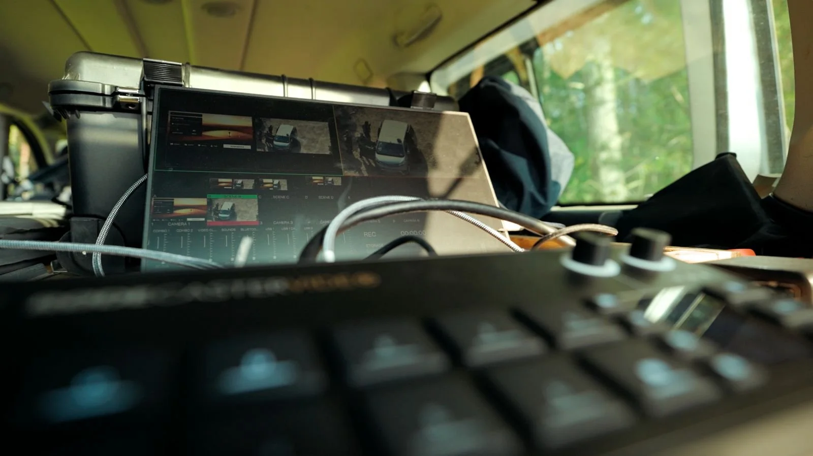 Innenraum eines Fahrzeugs mit einem Laptop, der Videobearbeitungssoftware zeigt, sowie einem Controller und diversen Kabeln. Das Fenster zeigt eine grüne Umgebung draußen.