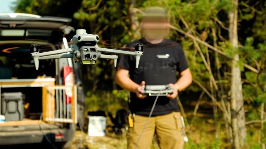 Drohne fliegt im Flug vor einem Mann mit Fernsteuerung in einem Wald.