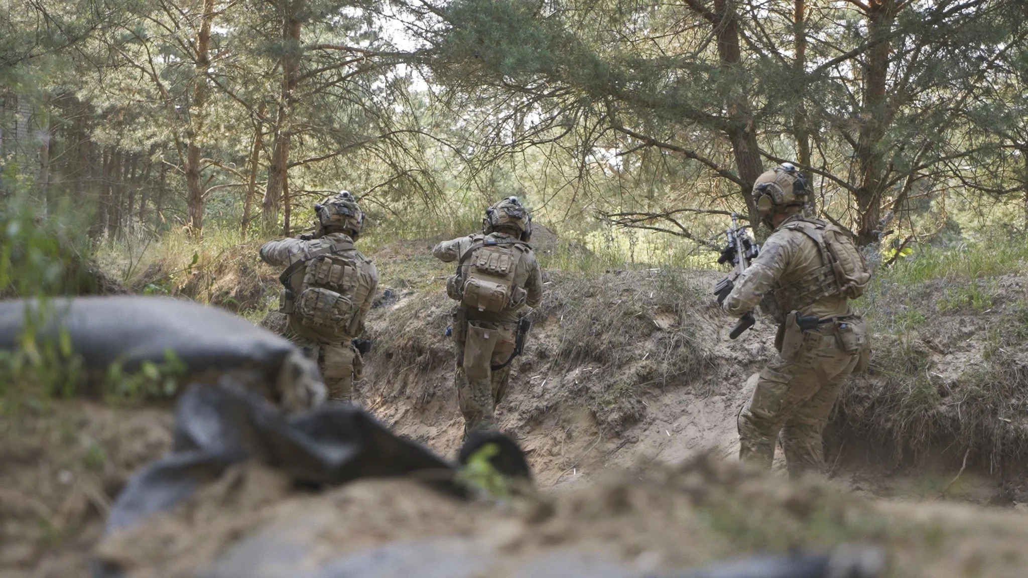 Vier Soldaten in Tarnuniform laufen durch den Wald mit Gewehren in Bereitschaft.