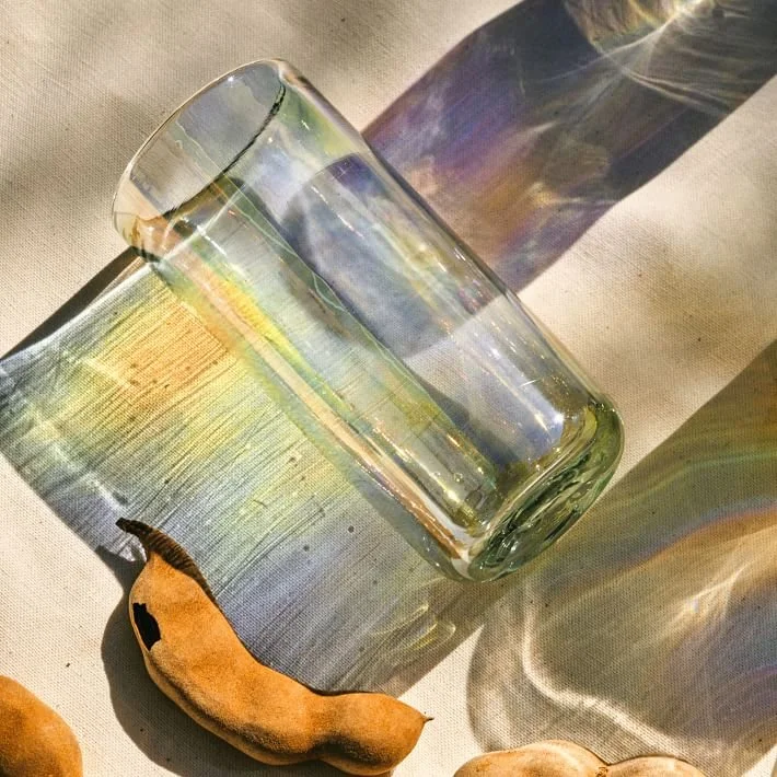 A clear glass tumbler lying on its side on a light-colored surface, with colorful light reflections on and cast by the glass. There are a few dried banana peels nearby.