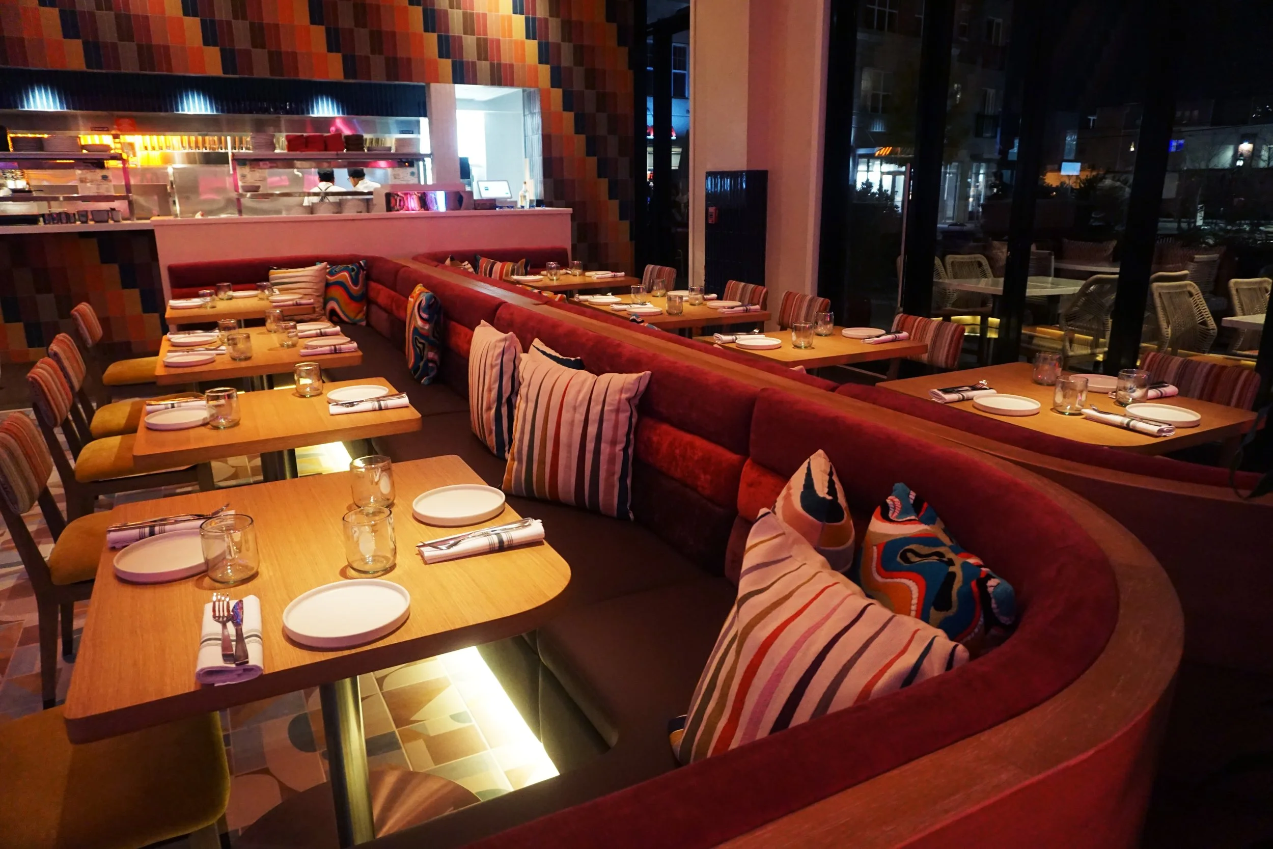 Interior of a restaurant with wooden tables set for dining, colorful striped and solid cushions on a curved red velvet banquette, and large windows showing outdoor seating at night.