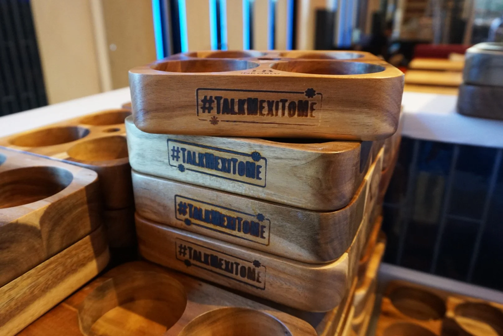 Stack of wooden cups or containers engraved with '#TALKMEXITOME' in black or blue ink, with other similar items nearby.