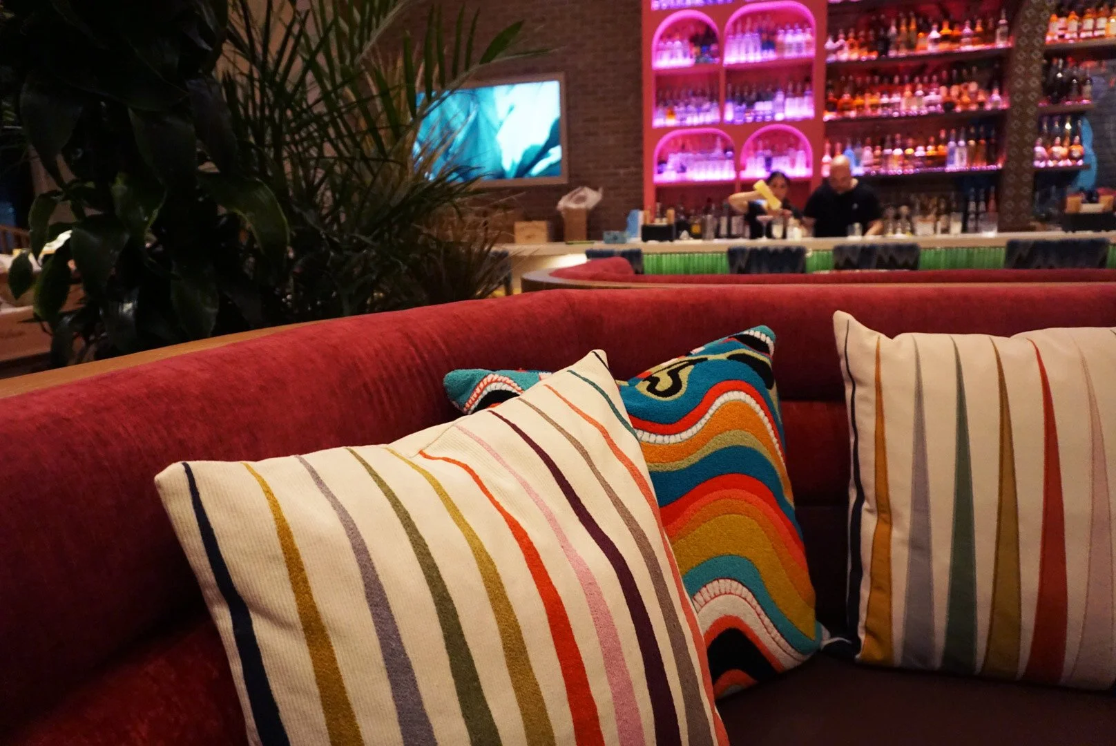 Colorful striped pillows on a red cushioned sofa in a lively bar or restaurant with a pink-lit bar in the background, and a plant partially visible on the left.