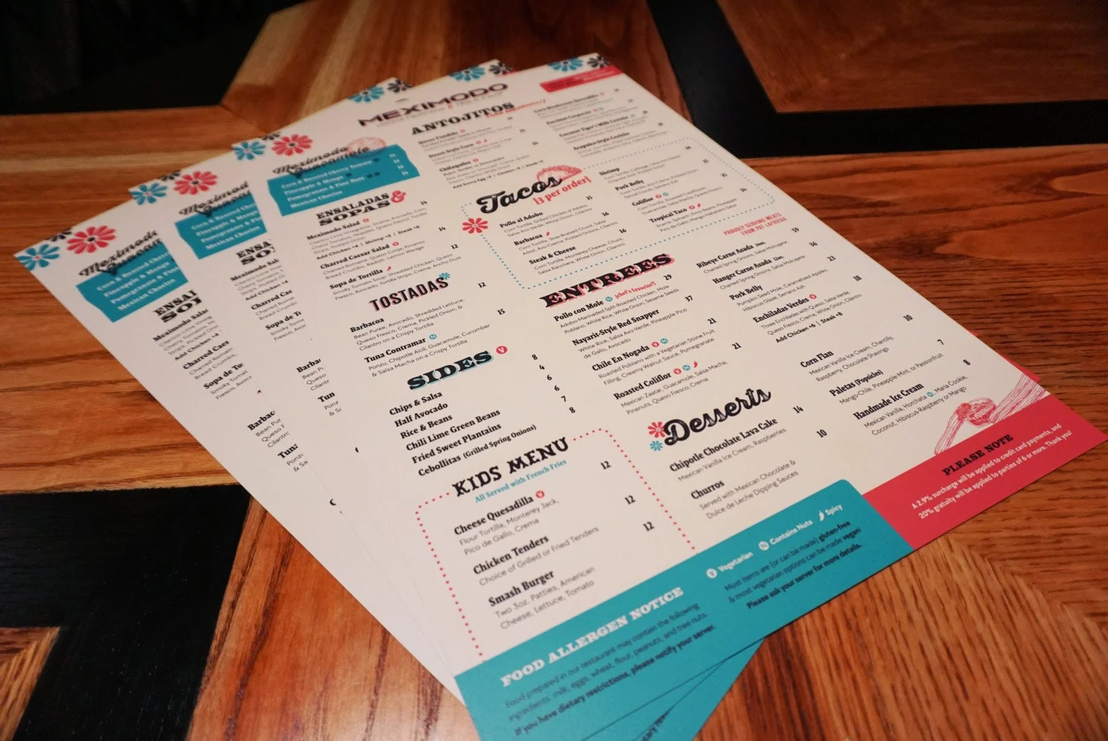 Meximundo restaurant menu on a wooden table with blue, red, and black text and decorative flowers.